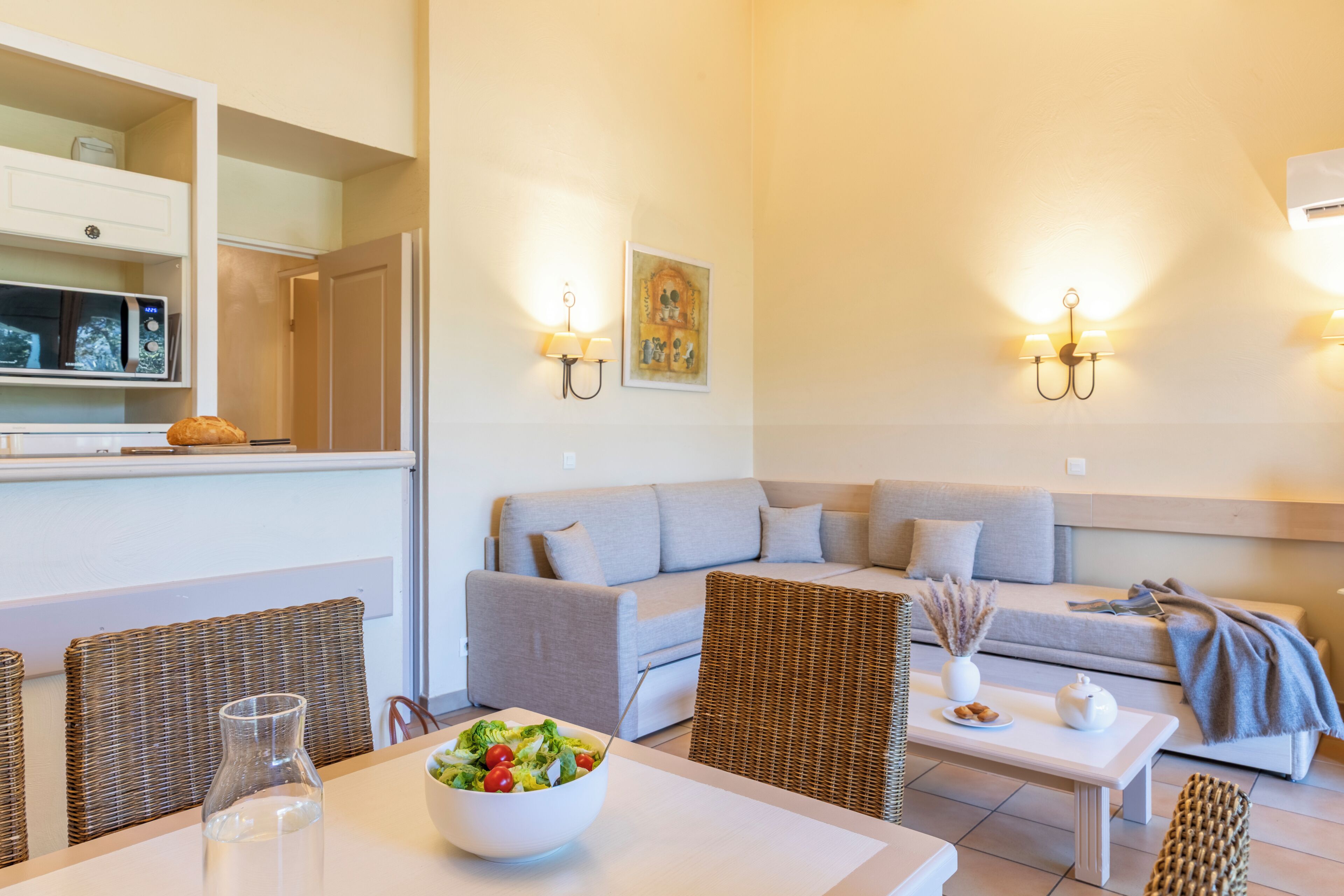 Een moderne woonkamer met een bank, rieten stoelen, een eettafel met een saladeschaal, wandlampen en een ingelijst kunstwerk.