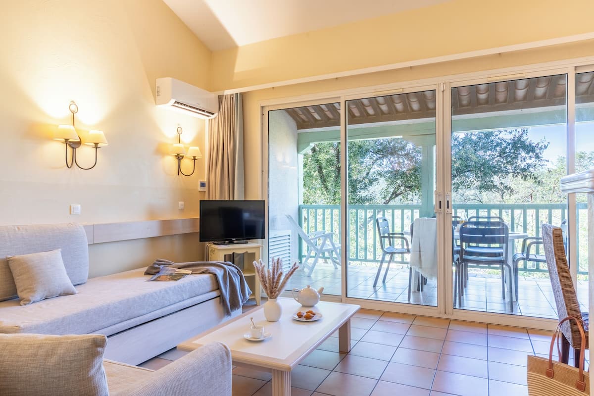 A modern living room with a sofa, a coffee table, and a TV. There is a sliding glass door leading to an outdoor patio with chairs and a table.