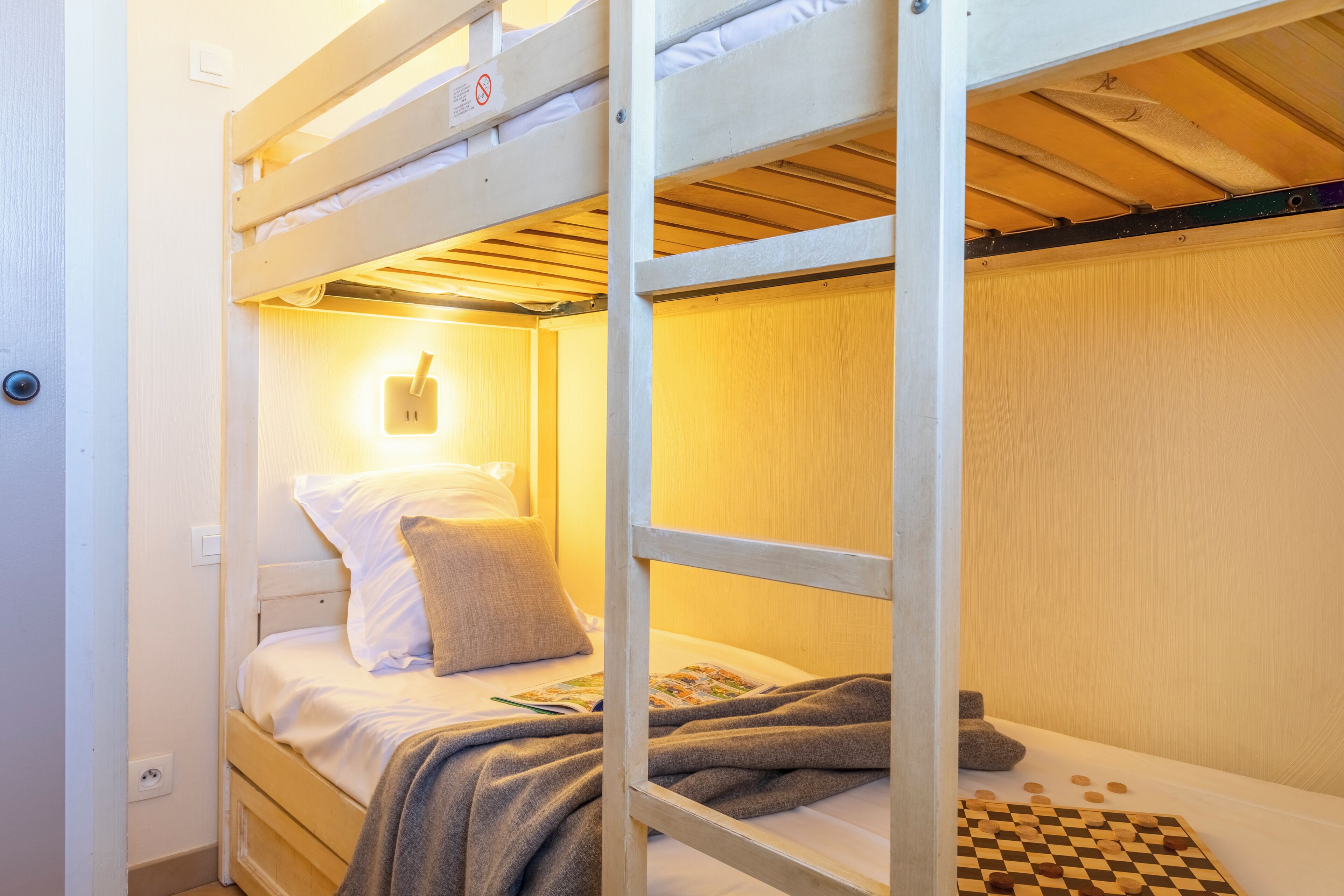 Una cama litera con un juego de mesa a cuadros, una lámpara de pared y un almohadón en la cama inferior.