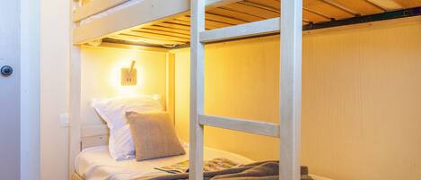 Una cama litera con un juego de mesa a cuadros, una lámpara de pared y un almohadón en la cama inferior.