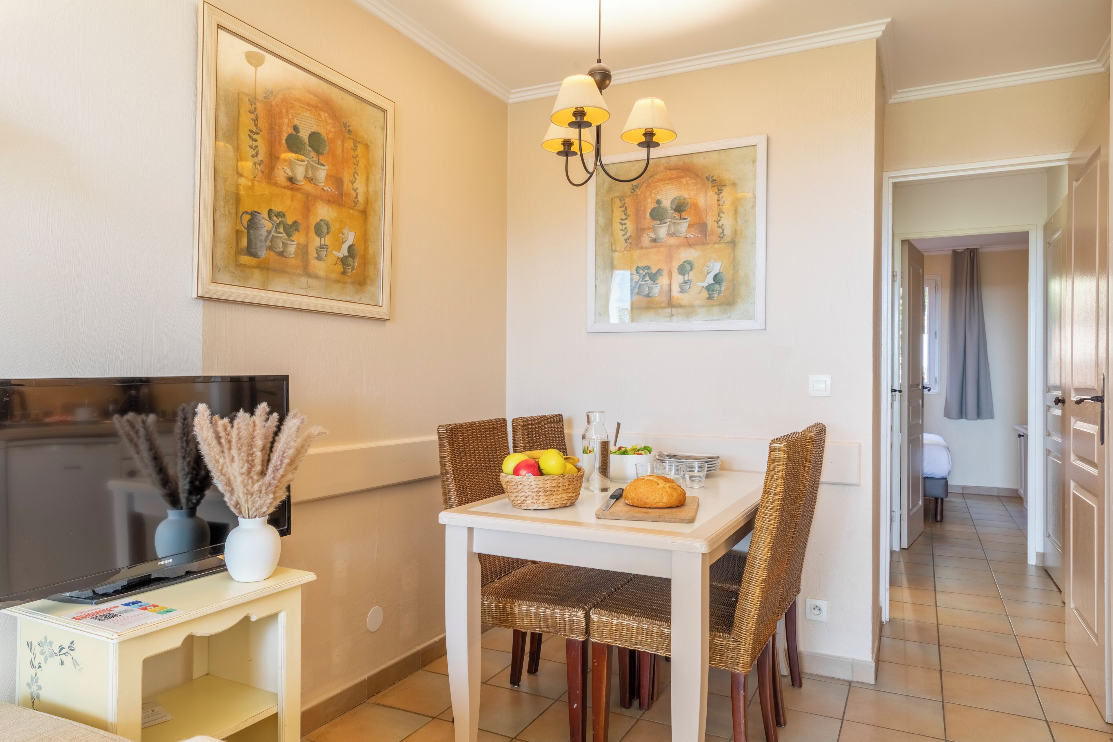 Un coin repas avec une table blanche, des chaises en osier, un téléviseur posé sur un meuble et des œuvres d’art accrochées aux murs.