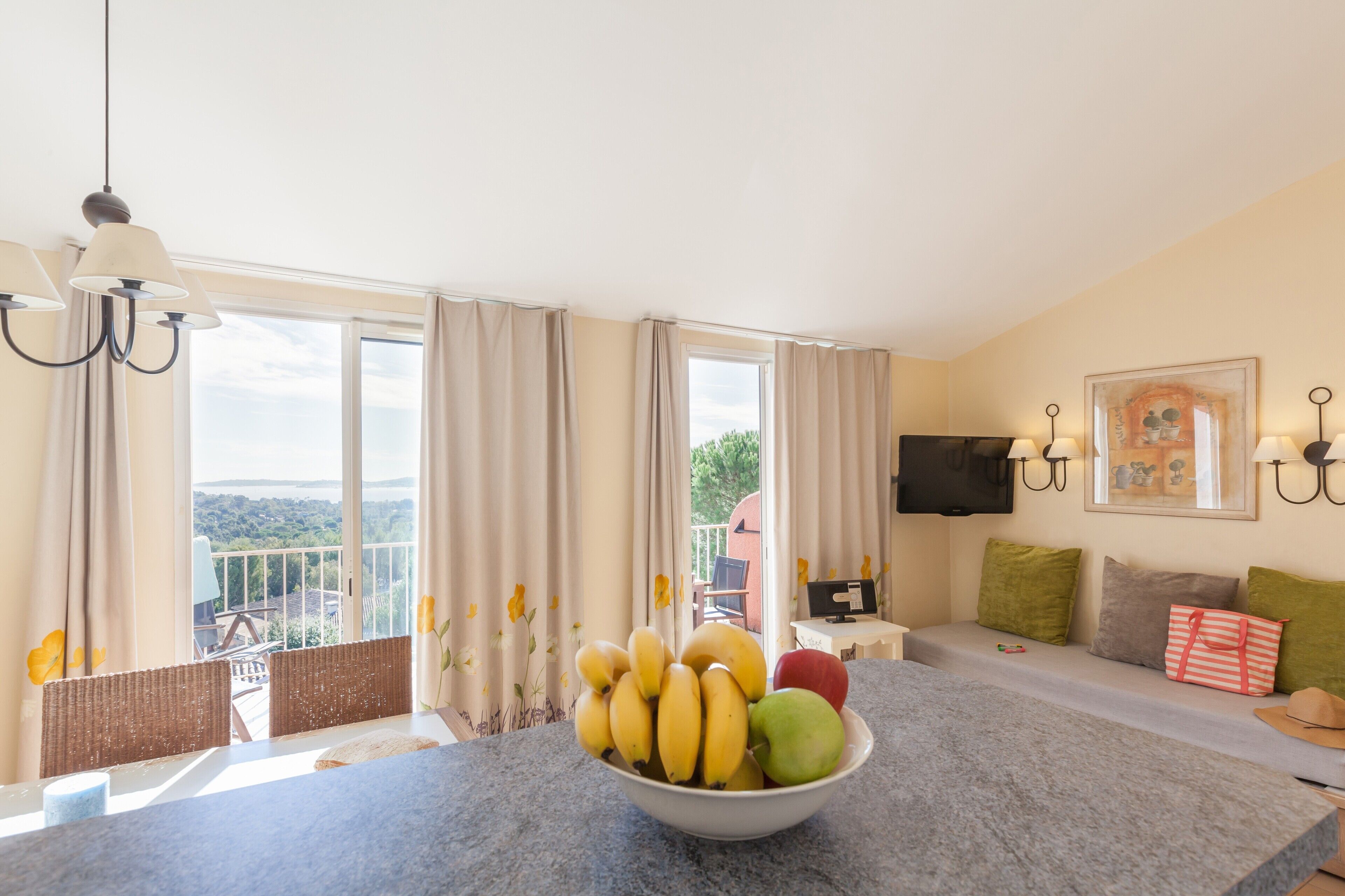 A room with a dining table, a bowl of fruit, a sofa, and a TV on the wall.