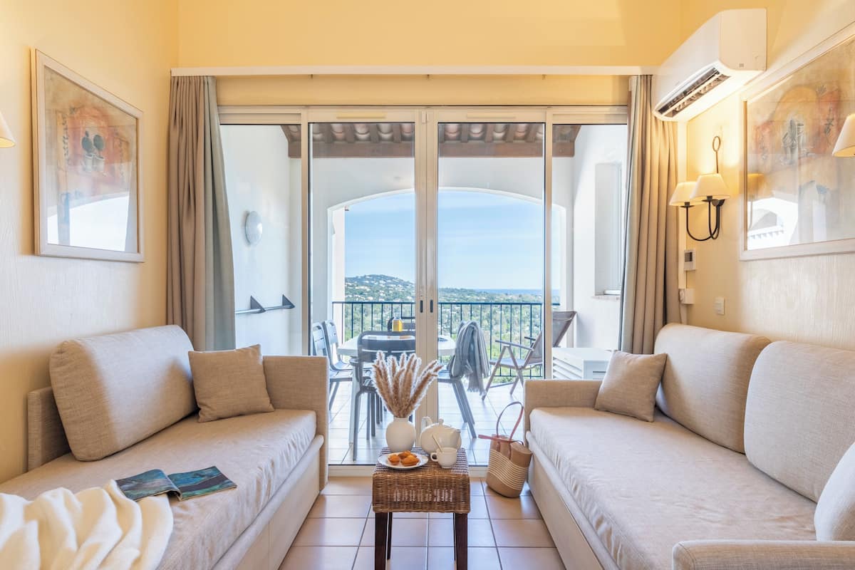 A hotel room with a balcony, two sofas, a small table with a vase and a plate of pastries, and a view of the city.