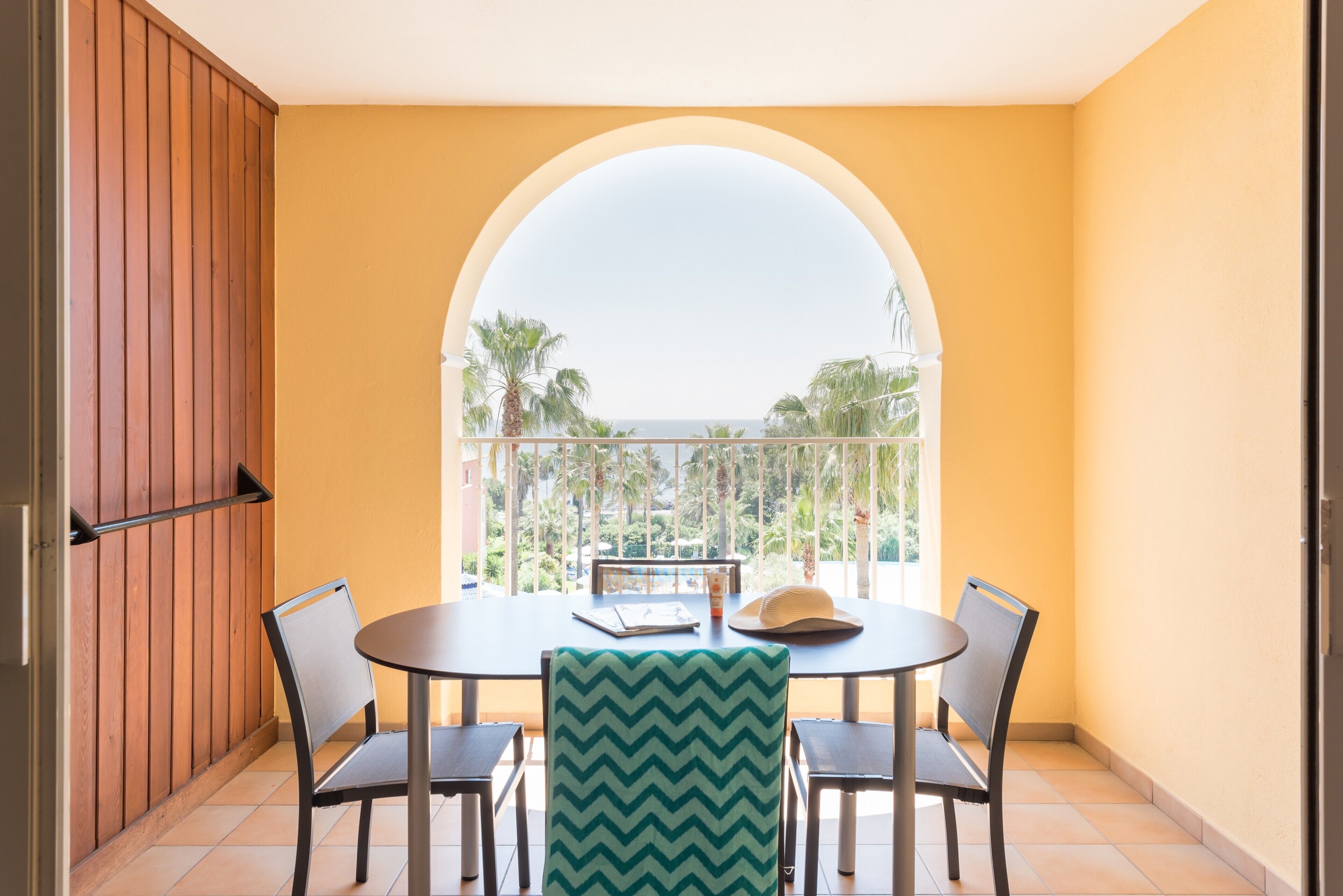 A balcony with a table and chairs, a view of palm trees, and a wooden wall.
