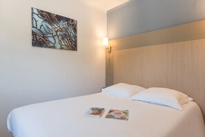 A neatly made bed with two pillows and two magazines on the bed. There is a wall-mounted light fixture above the bed and a piece of abstract art on the wall.