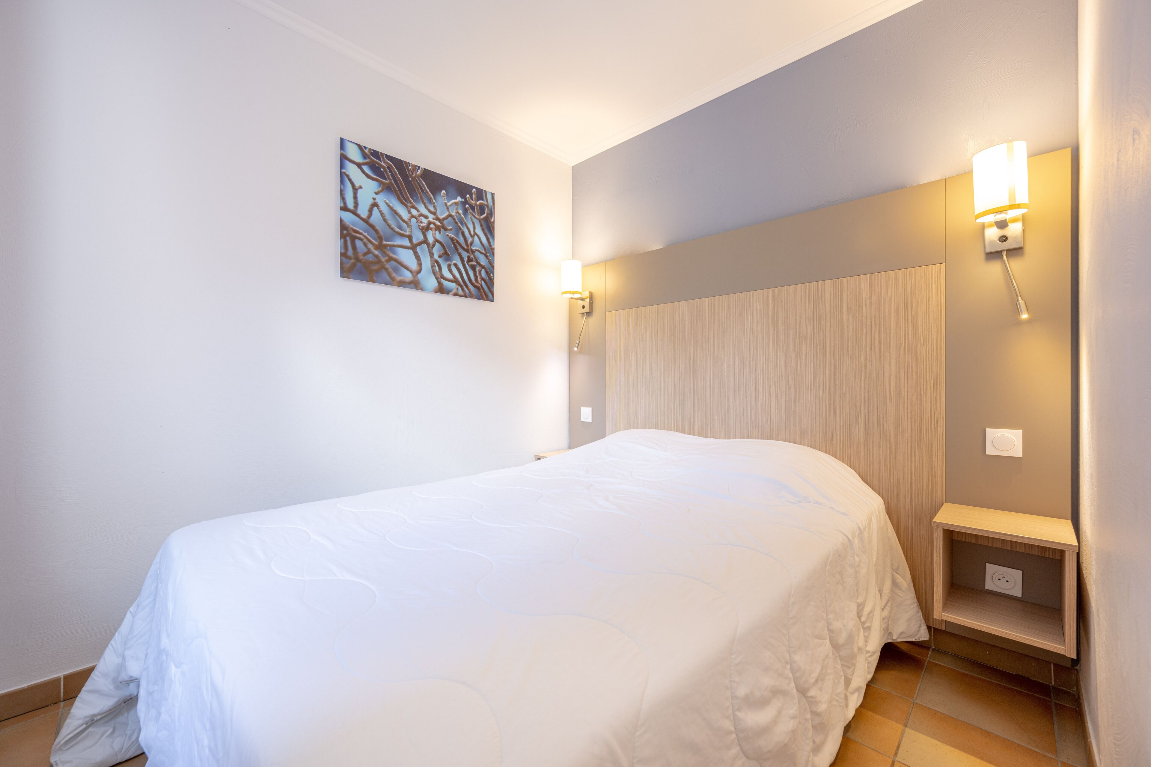 Un lit bien fait dans une chambre d’hôtel, avec un luminaire fixé au mur et une petite table de chevet.
