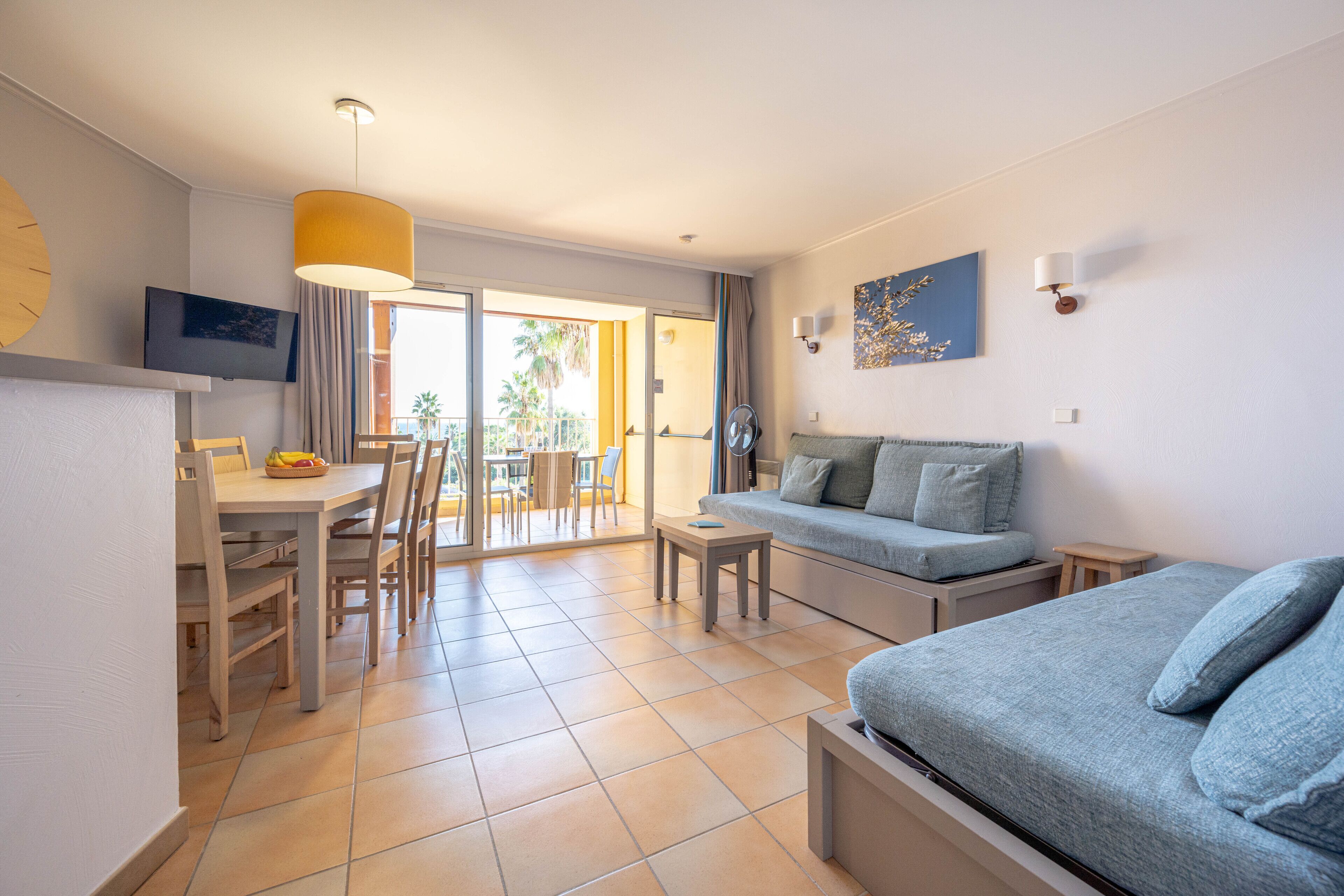 Una habitación de hotel moderna con cama, sofá, mesa de comedor con sillas, televisor y balcón con vista a palmeras.