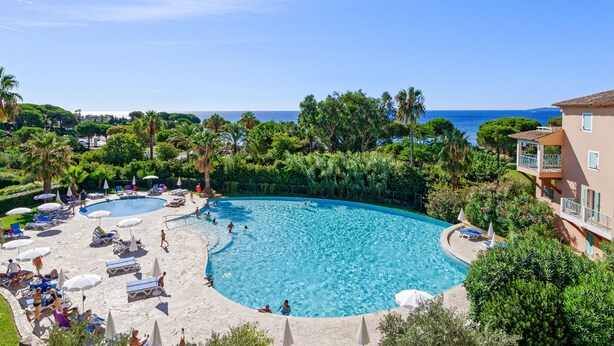 A large outdoor swimming pool surrounded by lounge chairs and umbrellas, with a view of the sea and lush greenery. - Pierre & Vacances Les Rivages des Issambres (Roquebrune-sur-Argens)