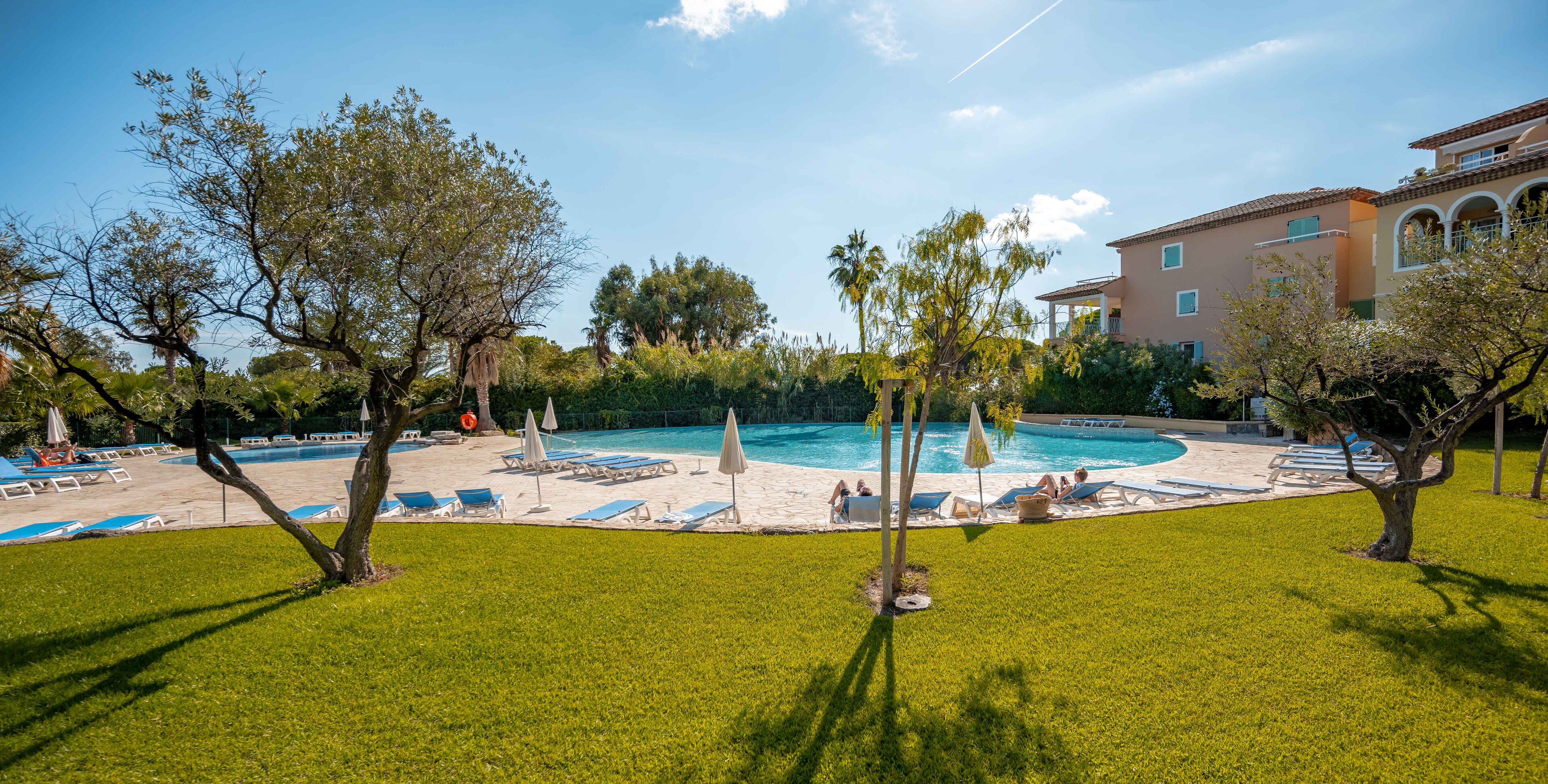 A pool area with lounge chairs and umbrellas, surrounded by trees and a building with arched windows.
