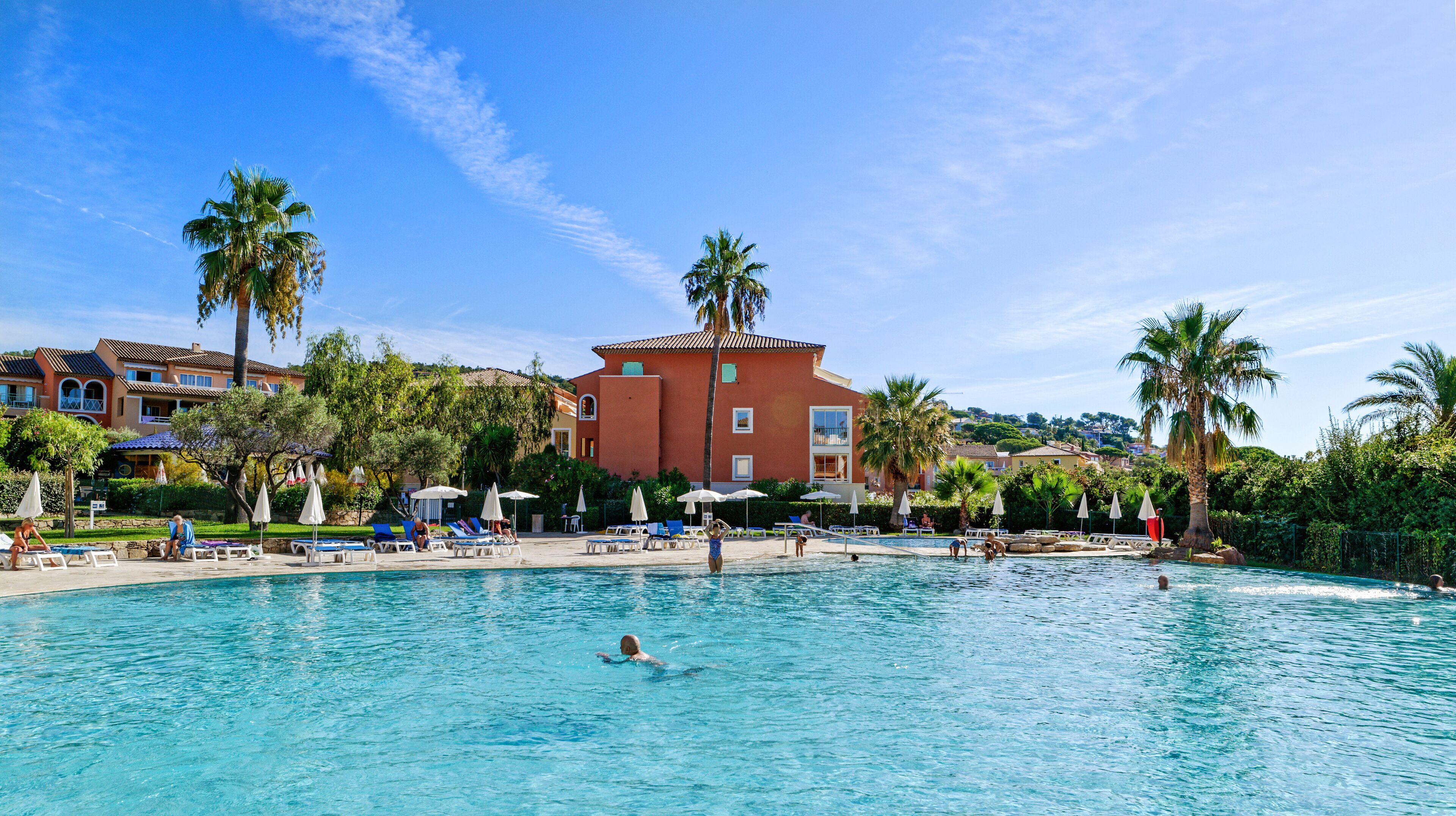 Una piscina con sillas de descanso y sombrillas, rodeada de palmeras y un edificio rojo con varias ventanas.