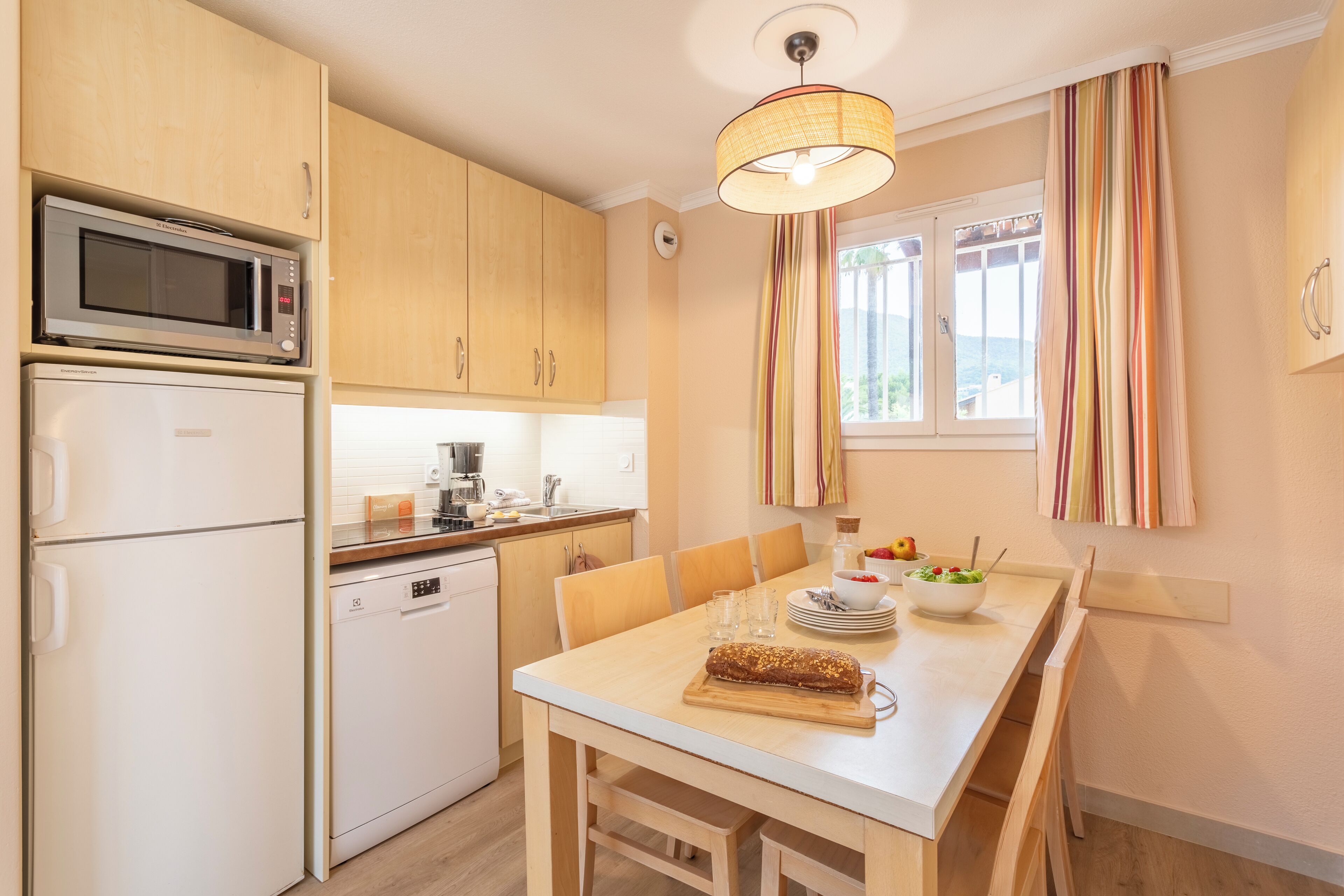 Una cocina moderna con microondas, refrigerador, lavavajillas y armarios de madera. Hay una mesa de comedor con sillas, una lámpara colgante y una ventana con cortinas.