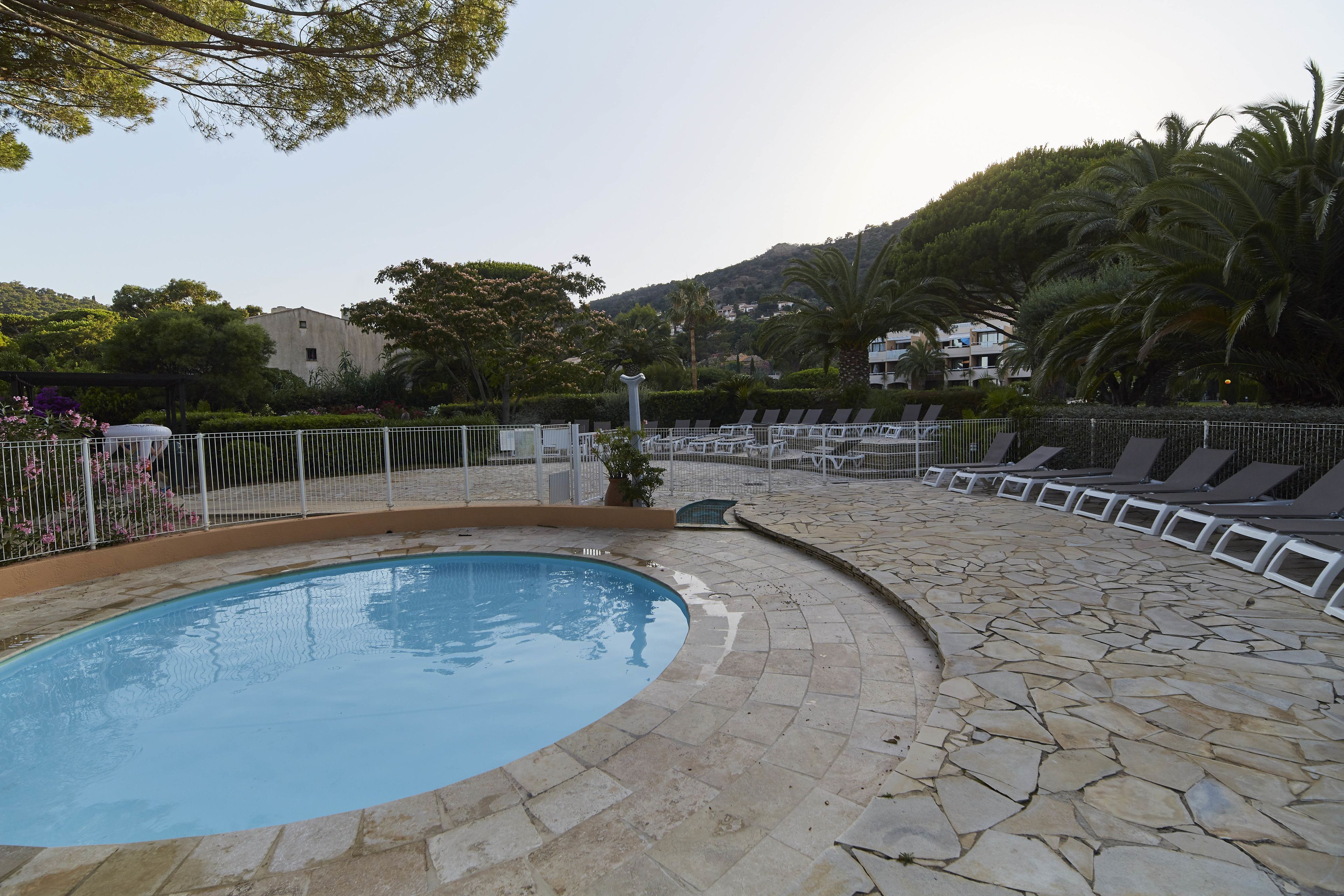 A swimming pool with a stone deck, lounge chairs, and a view of a hill with buildings and trees.