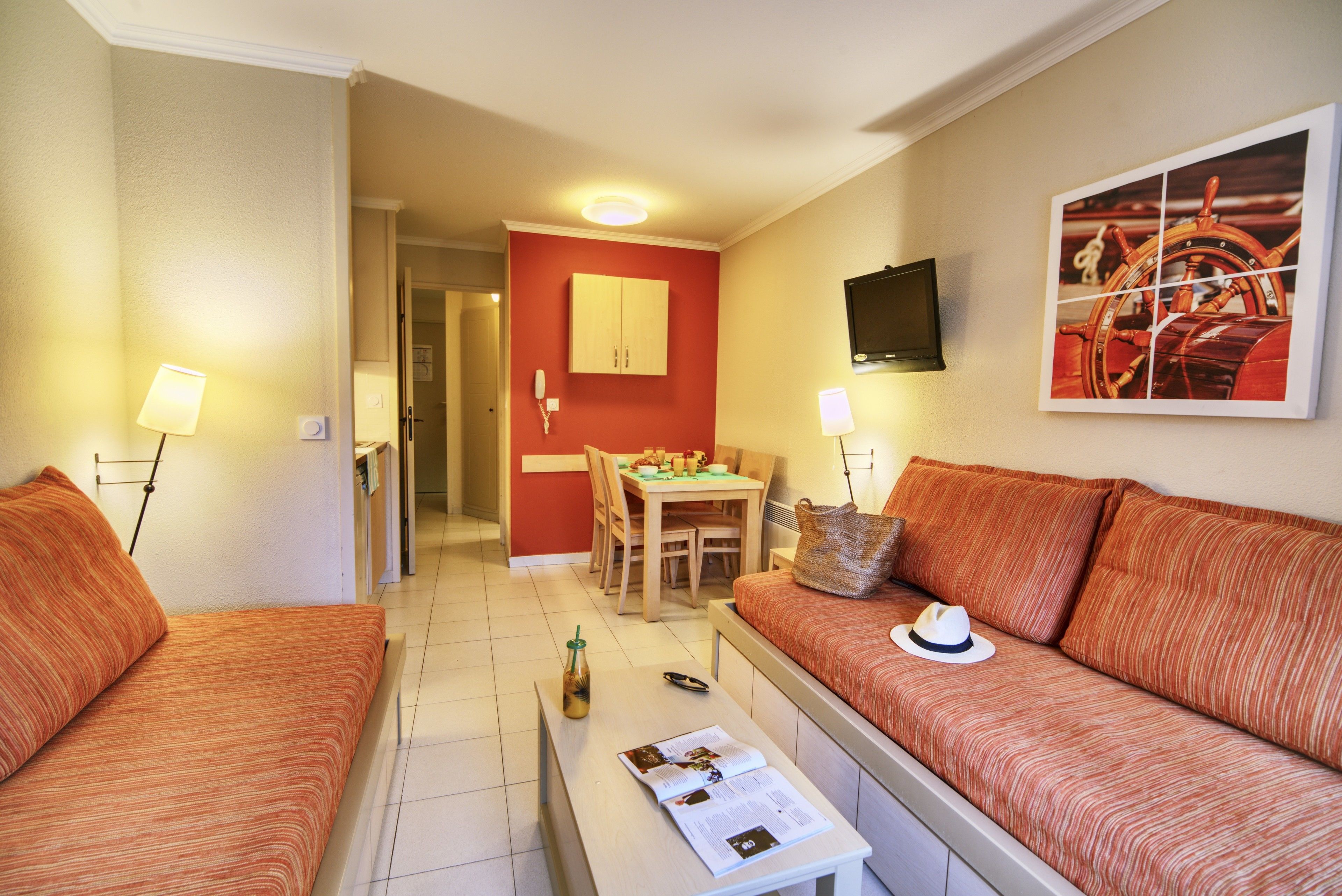 Une chambre d’hôtel avec deux canapés à rayures orange, une petite table avec un magazine, un coin repas avec une table et des chaises, un mur d’accent rouge et un téléviseur fixé au mur.