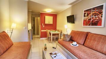 Une chambre d’hôtel avec deux canapés à rayures orange, une petite table avec un magazine, un coin repas avec une table et des chaises, un mur d’accent rouge et un téléviseur fixé au mur.