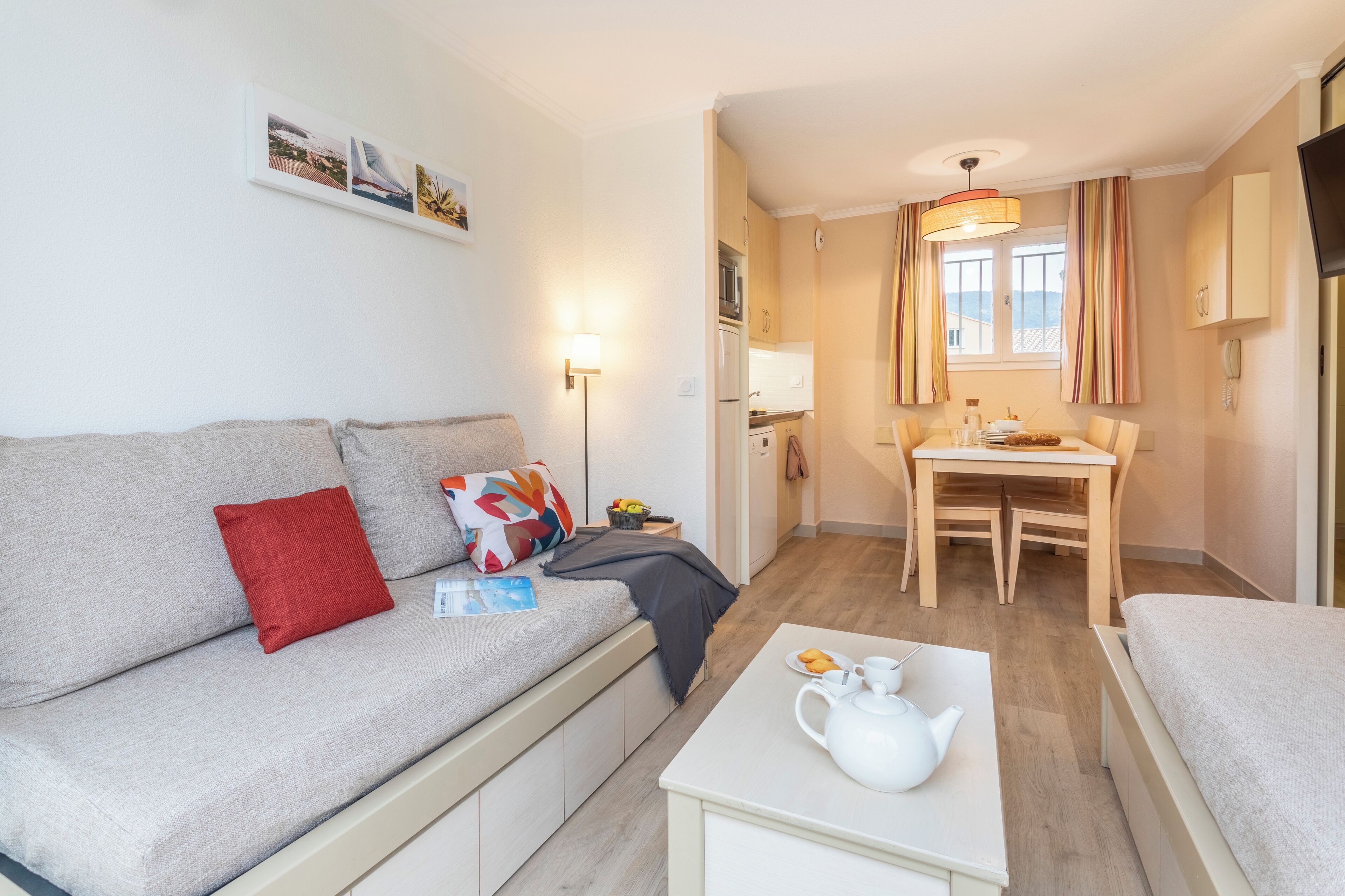 A compact living space with a sofa, a small table with a teapot, a dining table, and a kitchen area.