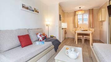 A compact living space with a sofa, a small table with a teapot, a dining table, and a kitchen area.