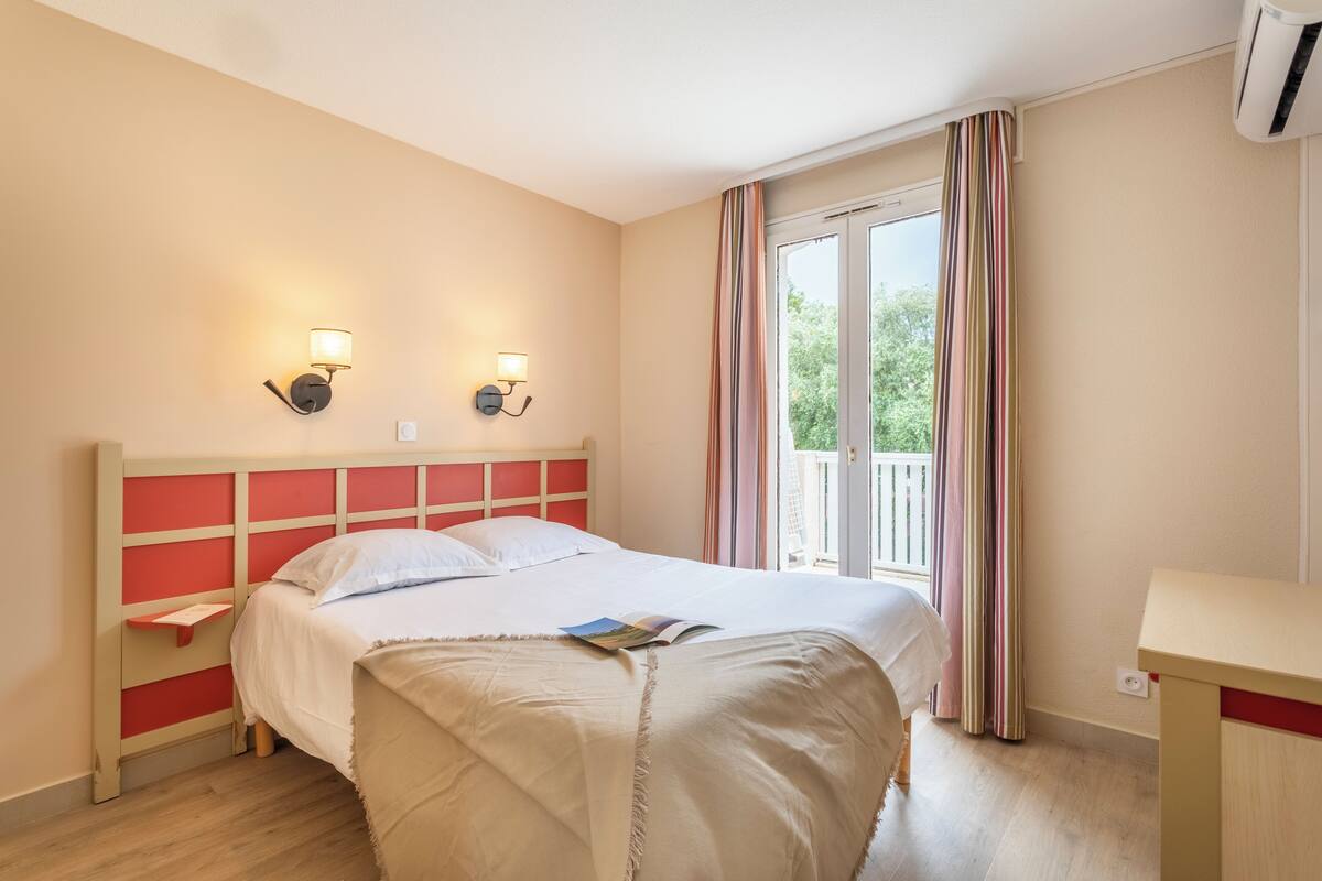 Ein Hotelzimmer mit einem Bett, Nachttischlampen, einem Fenster mit Vorhängen und einem kleinen Tisch.