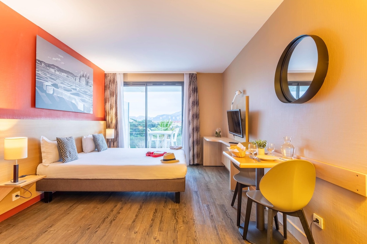 Une chambre d’hôtel équipée d’un lit, d’un bureau, d’une chaise, d’une télévision et d’une grande fenêtre donnant sur un beau paysage.