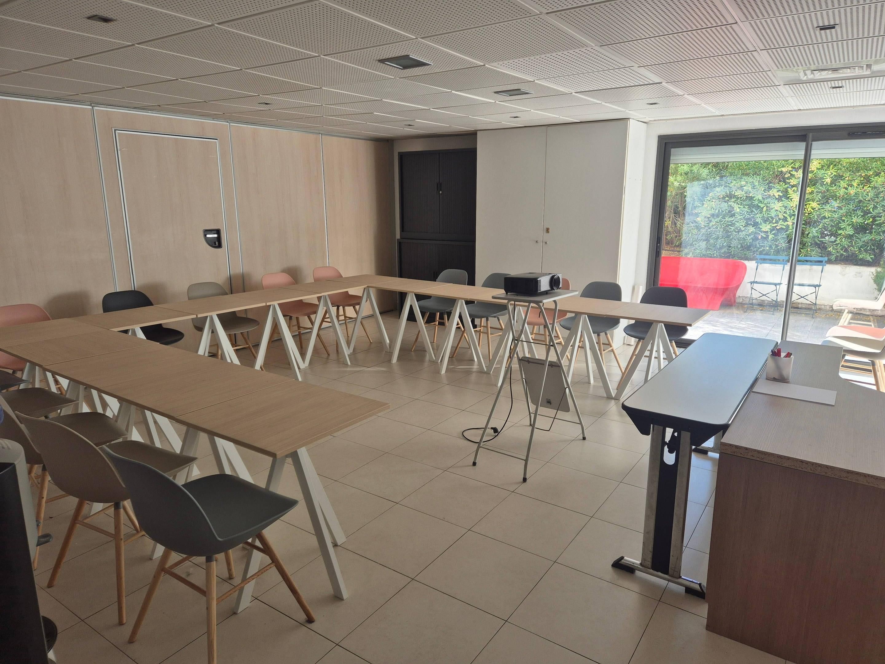 Una sala de conferències amb una taula llarga, cadires, un projector i una finestra amb vista a vegetació.