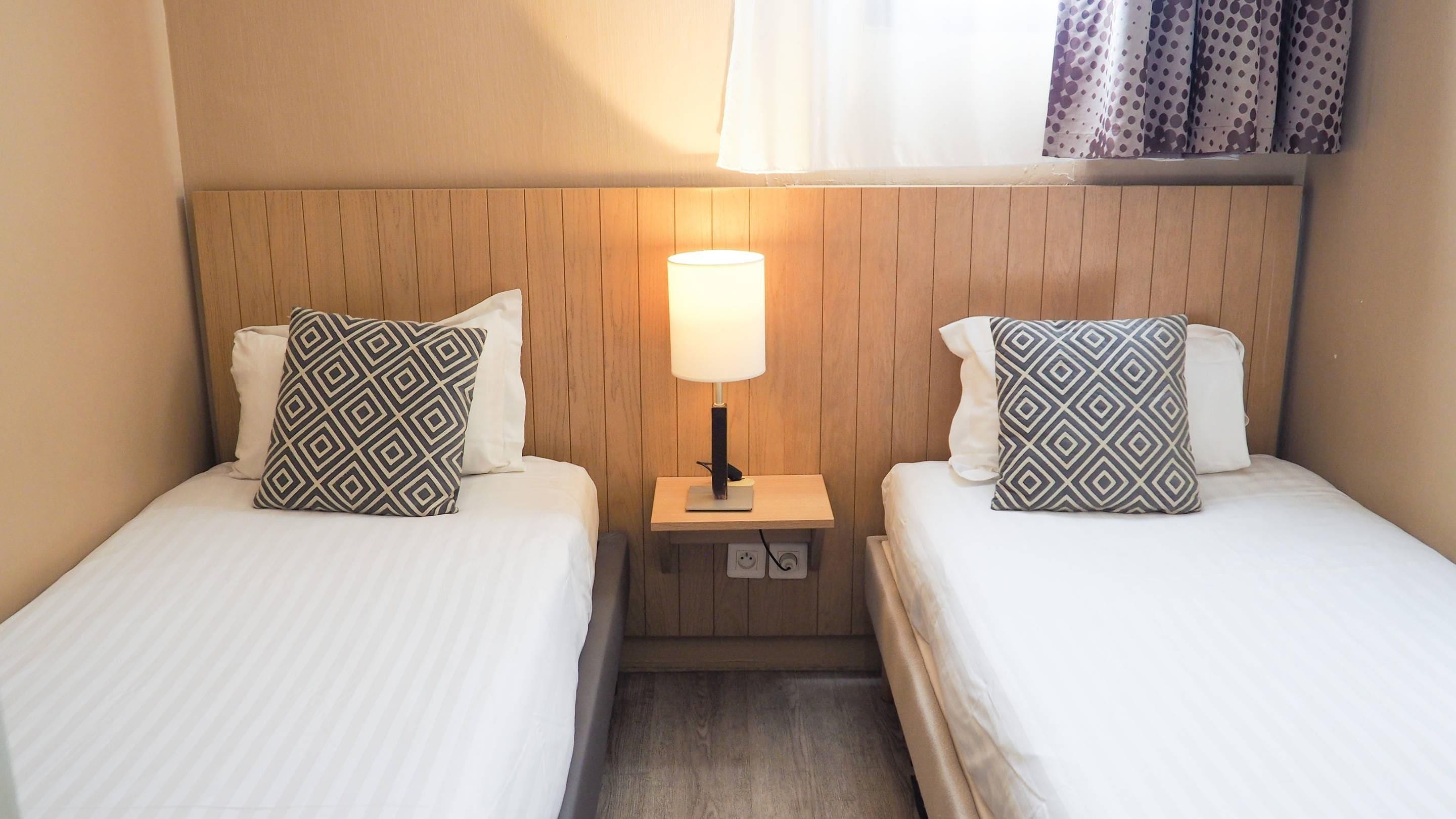 A hotel room with two beds, each with patterned pillows, a bedside table with a lamp, and a wooden headboard.