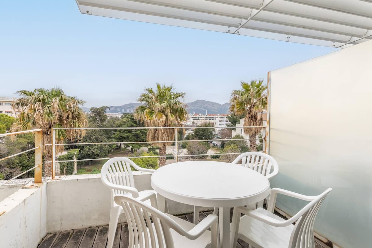 Un balcon avec une table ronde et des chaises blanches, donnant sur un paysage urbain avec des palmiers et des immeubles.
