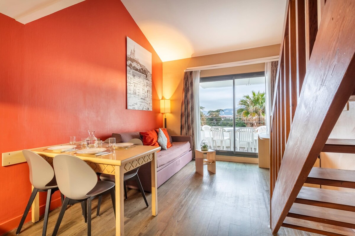 Een kamer met een eettafel, stoelen, een bank, een lamp en uitzicht door een groot raam.
