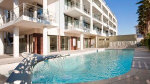 A modern hotel with a swimming pool, multiple balconies, and a clear blue sky.