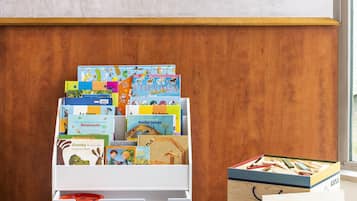 A room with a white storage box labeled "PETIT BAZAR" filled with children's books, a wooden cabinet with a "KAPIA" sign, and a blue carpet with a world map design.