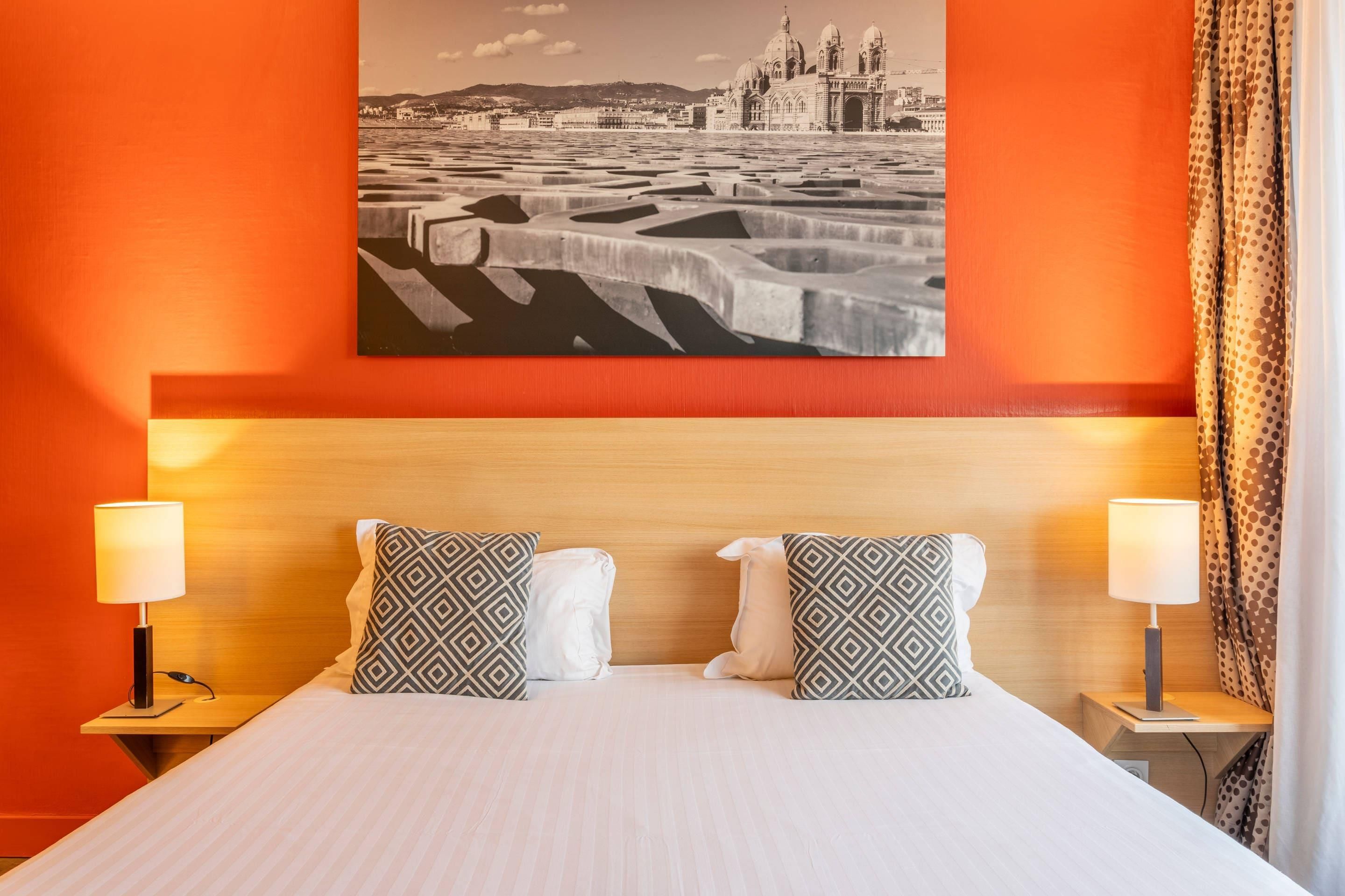 A hotel room with a large bed, two nightstands with lamps, patterned pillows, and a cityscape painting on the wall.