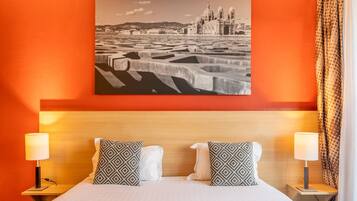 A hotel room with a large bed, two nightstands with lamps, patterned pillows, and a cityscape painting on the wall.