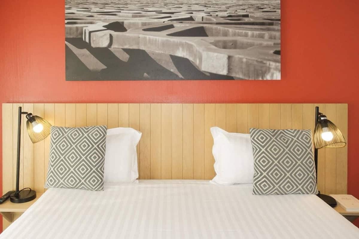 Une chambre d’hôtel avec une tête de lit en bois, deux coussins à motifs, un couvre-lit blanc et deux lampes de chevet.