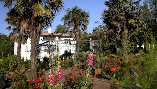 A white building with a red roof, surrounded by palm trees and flowering plants. - Pierre & Vacances Residence Le Domaine de Bordaberry (Urrugne)