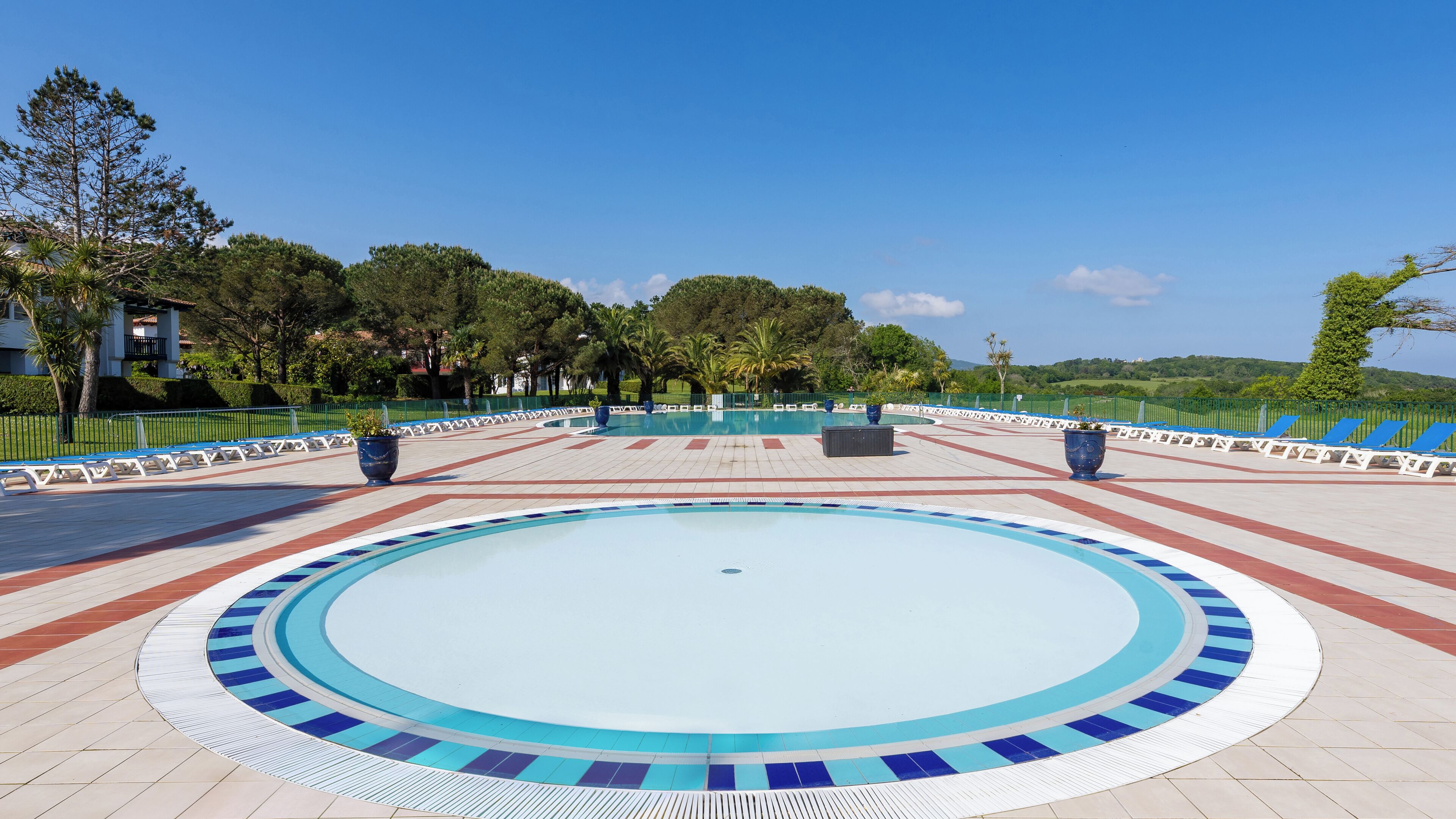 Una piscina circular con borde de azulejos, rodeada por una zona pavimentada con bancos y plantas en macetas.