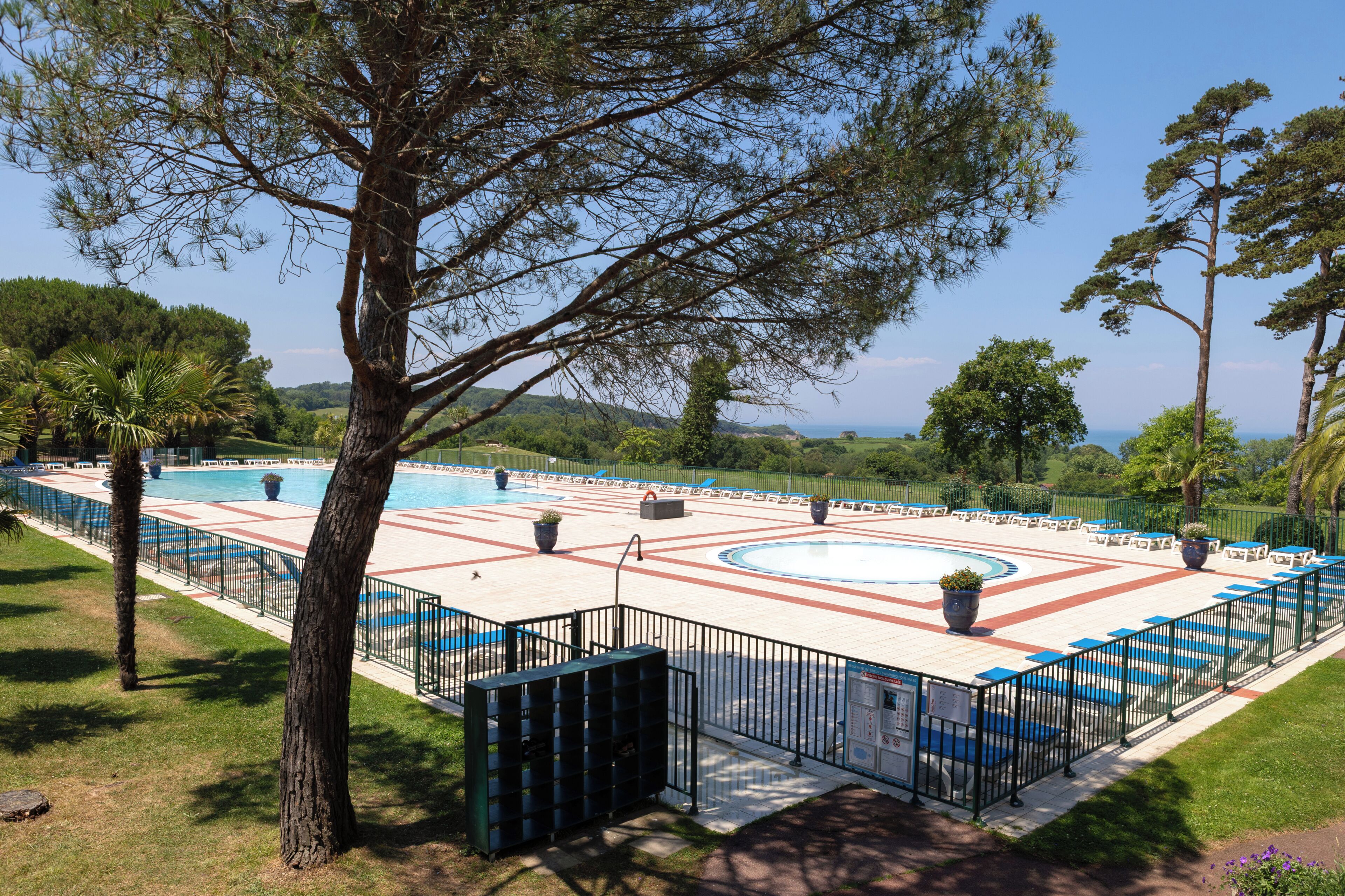 En swimmingpool med geometrisk flisemønster, omgivet af et hegn, og træer i baggrunden.
