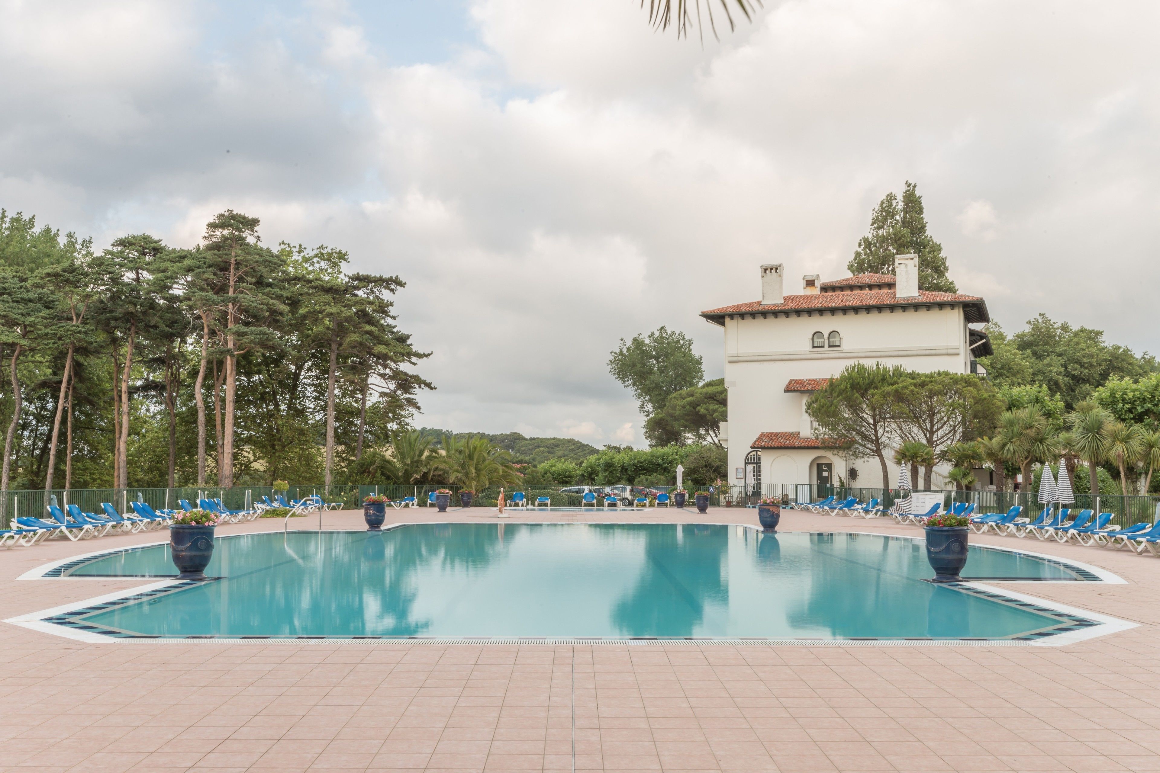 A swimming pool with lounge chairs, a white building, and trees in the background.