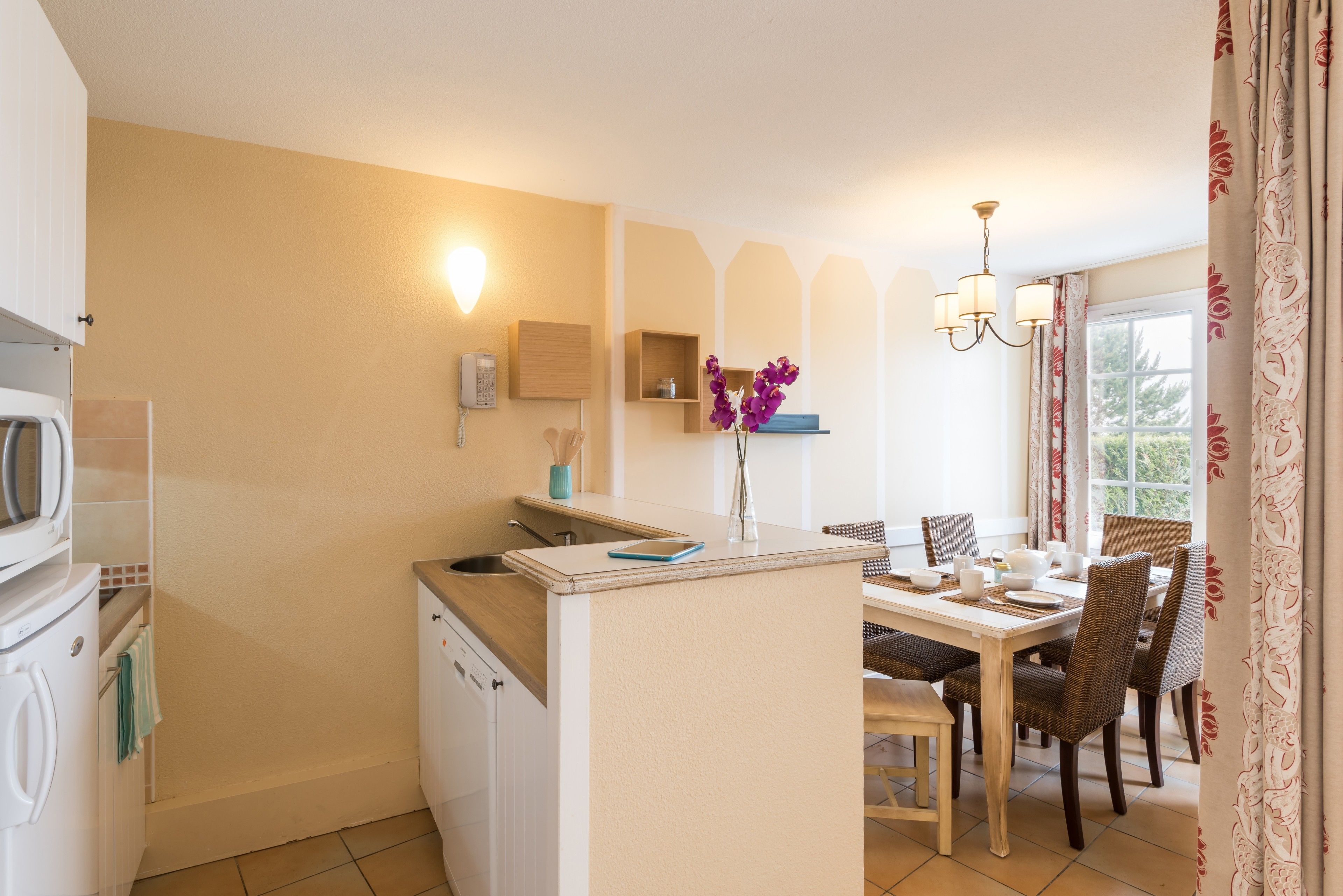 Una cocina funcional con microondas, cafetera y refrigerador pequeño. Hay un comedor con una mesa dispuesta para cuatro personas, rodeada de sillas de mimbre. Un florero con flores moradas está sobre la encimera, y un candelabro cuelga sobre la mesa de comedor.