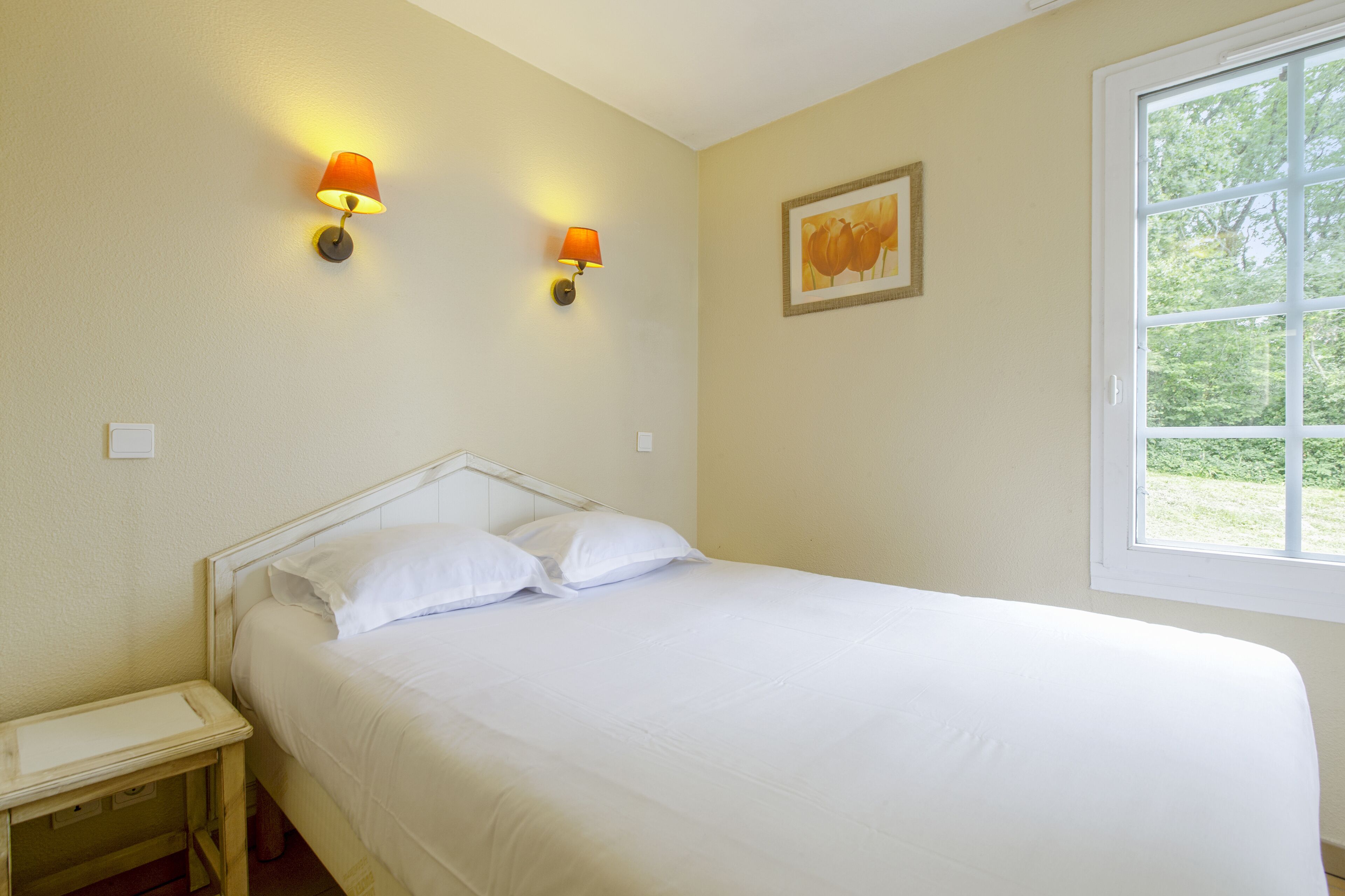 Un dormitorio con una cama, dos lámparas fijadas a la pared, una ventana y un cuadro enmarcado.