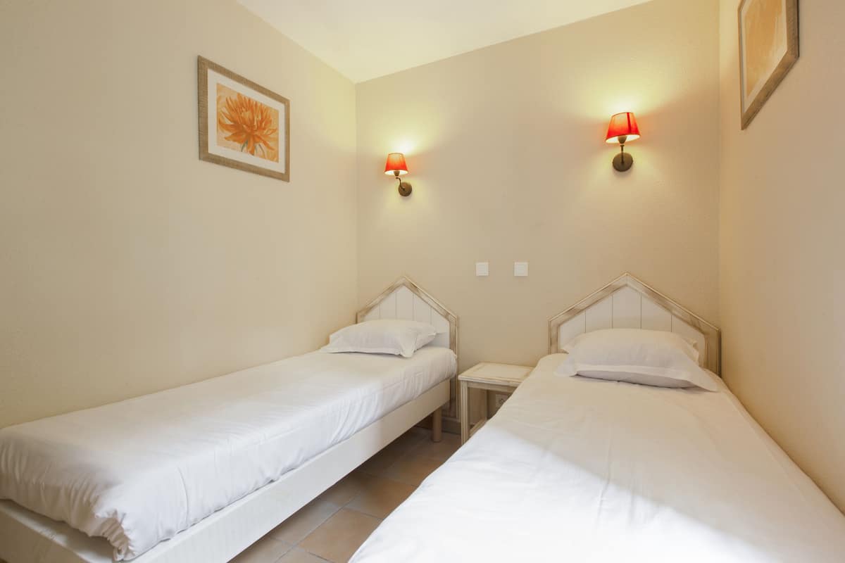 A room with two single beds, each with a wooden headboard and white bedding. There are two wall-mounted lamps with red shades.