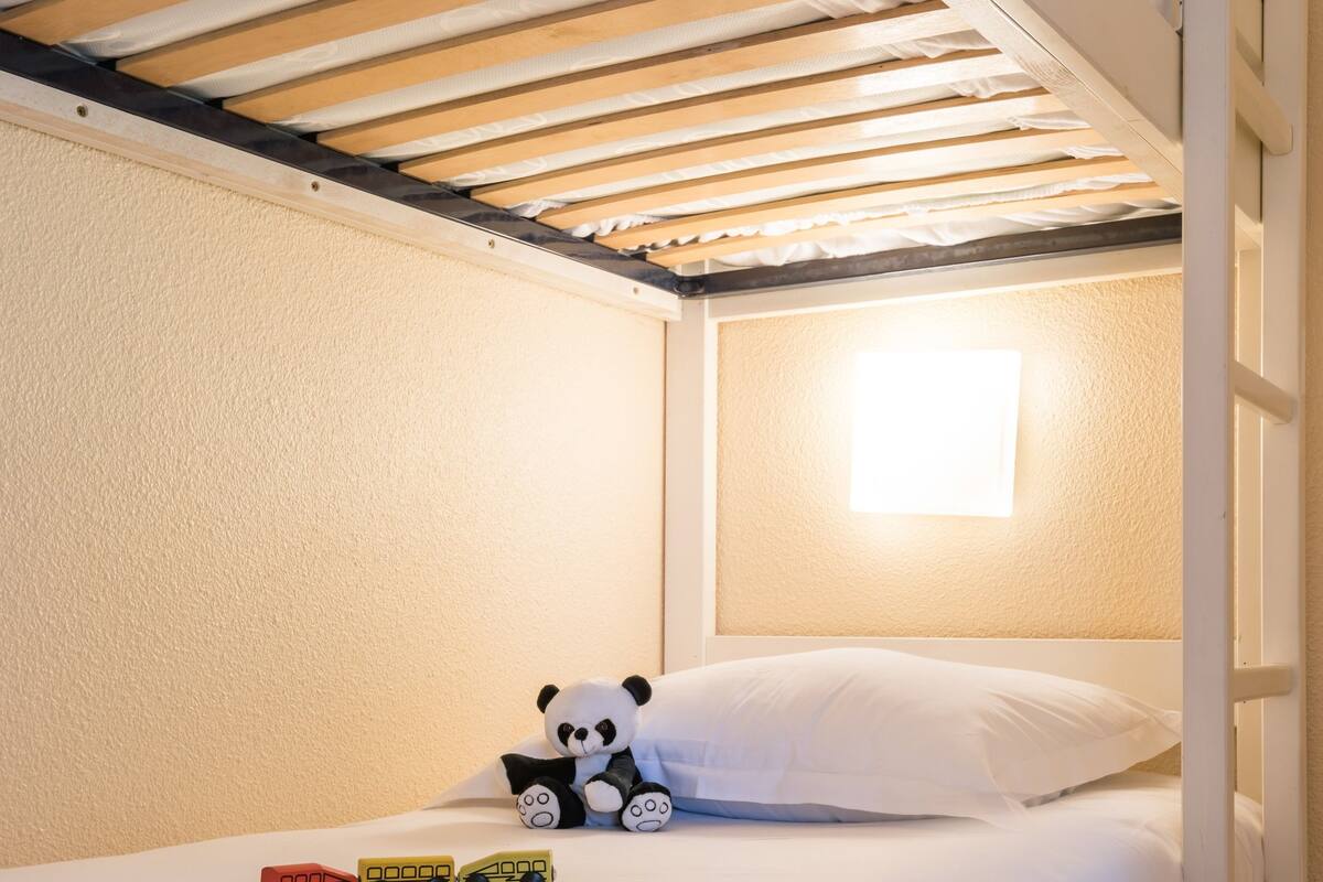A bunk bed with a white mattress, a small bedside table, a lamp, and a stuffed panda toy.