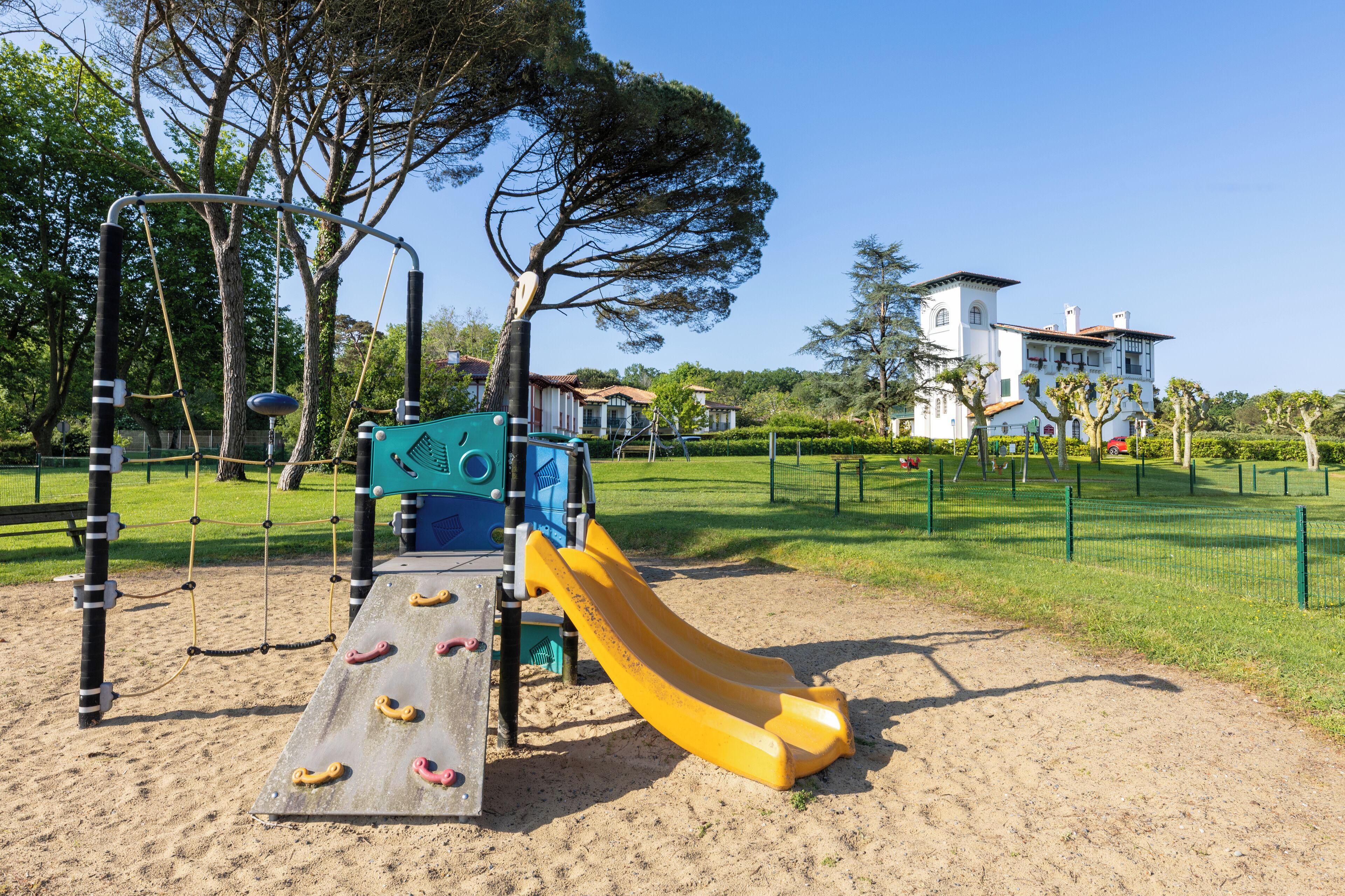 Un parque infantil con tobogán y estructuras para escalar, rodeado por una zona vallada y árboles.