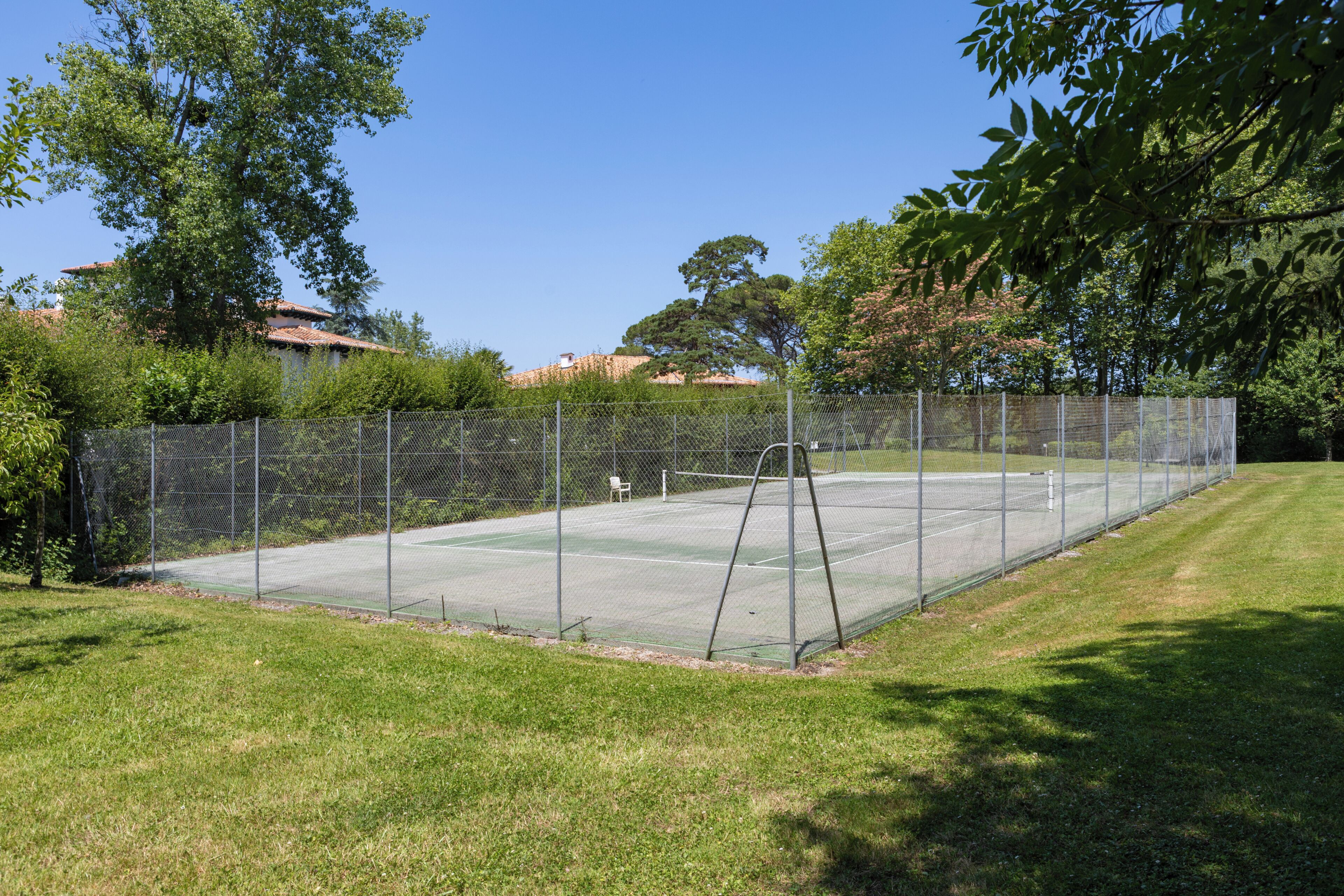 A tennis court surrounded by a chain-link fence, with a grassy area in the foreground and trees in the background.