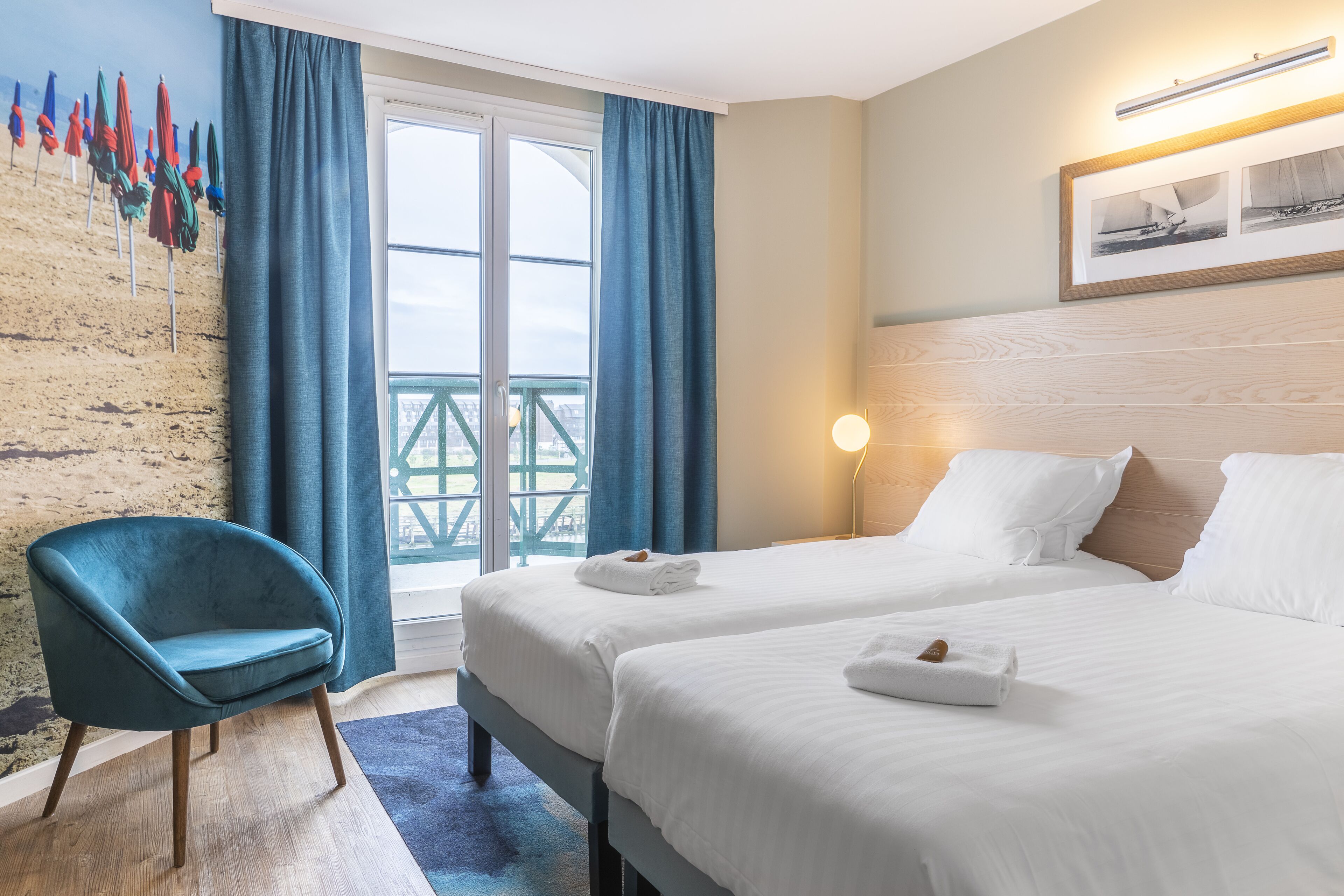 Habitación de hotel con dos camas, un sillón verde azulado, un ventanal con cortinas y un cuadro colgado en la pared.