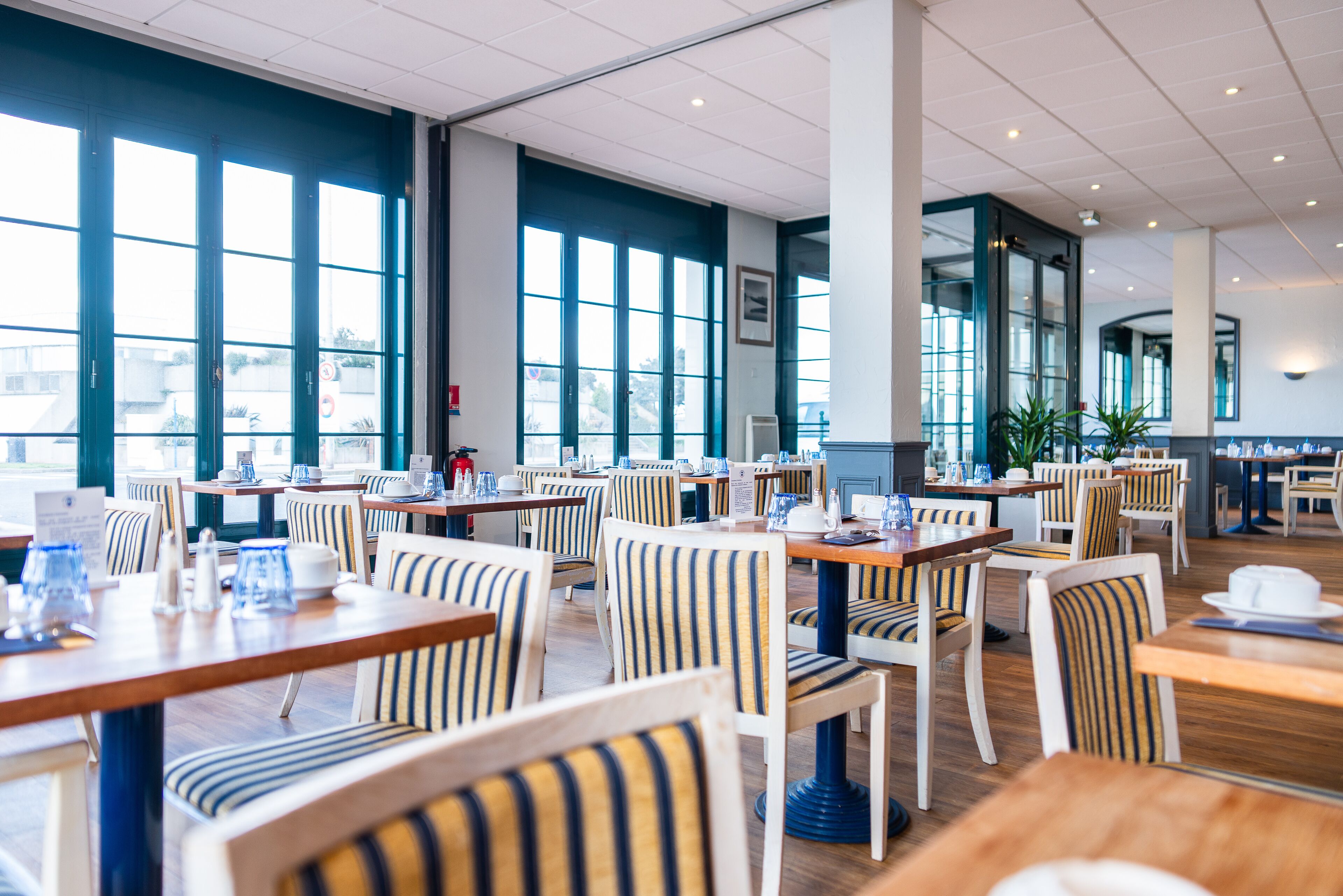 Un restaurante espacioso con techos altos, ventanas amplias y sillas a rayas. Las mesas están dispuestas con platos, vasos y cubiertos.