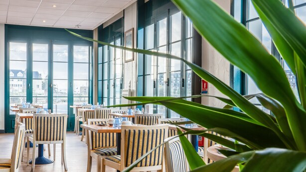 En restaurant med træborde og stribede stole, store vinduer og udsigt til bygninger udefra.