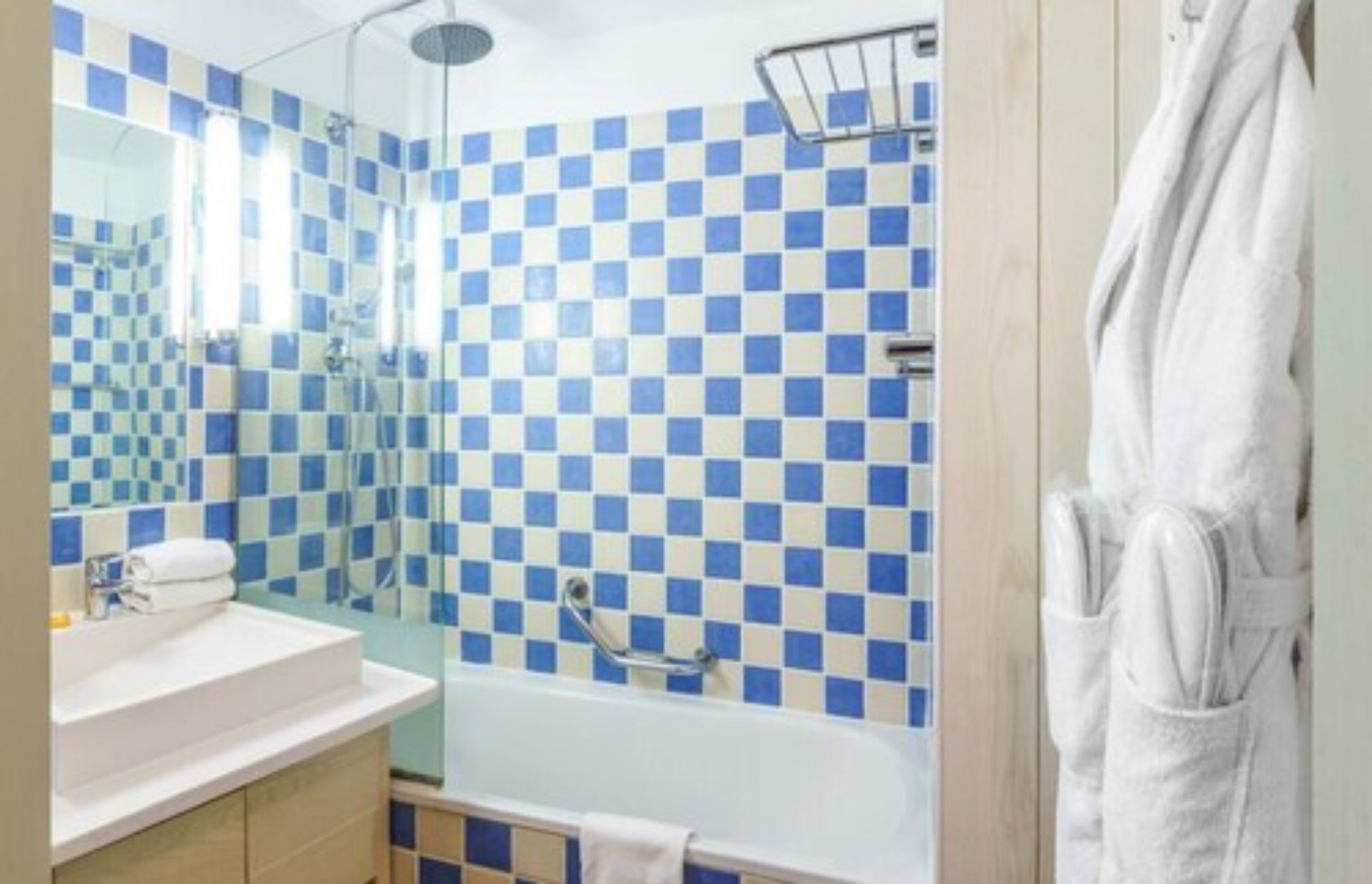 Ein Badezimmer mit Wand aus blau-weißen Schachfeldern, einer weißen Badewanne, einer Duschkabine und einem weißen Bademantel, der an der Wand hängt.