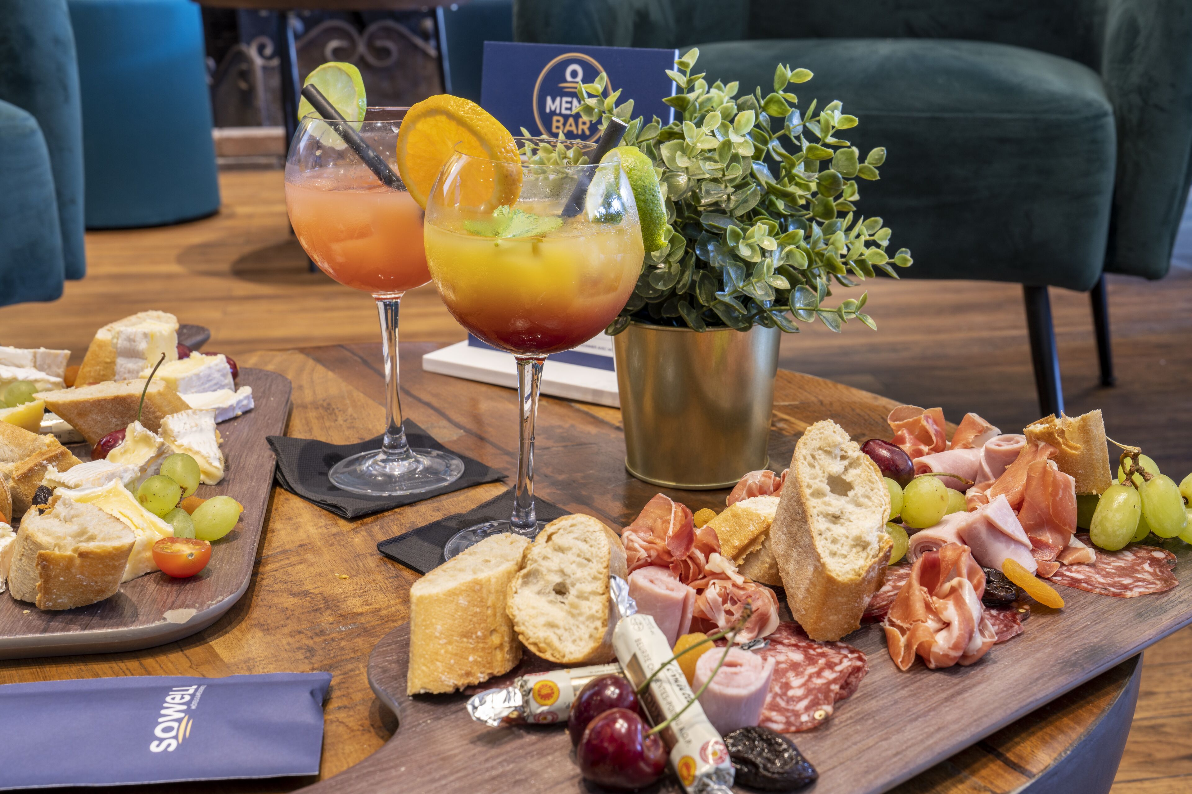 Una tabla de fiambres con diversos quesos, carnes y frutas, acompañada de dos bebidas.