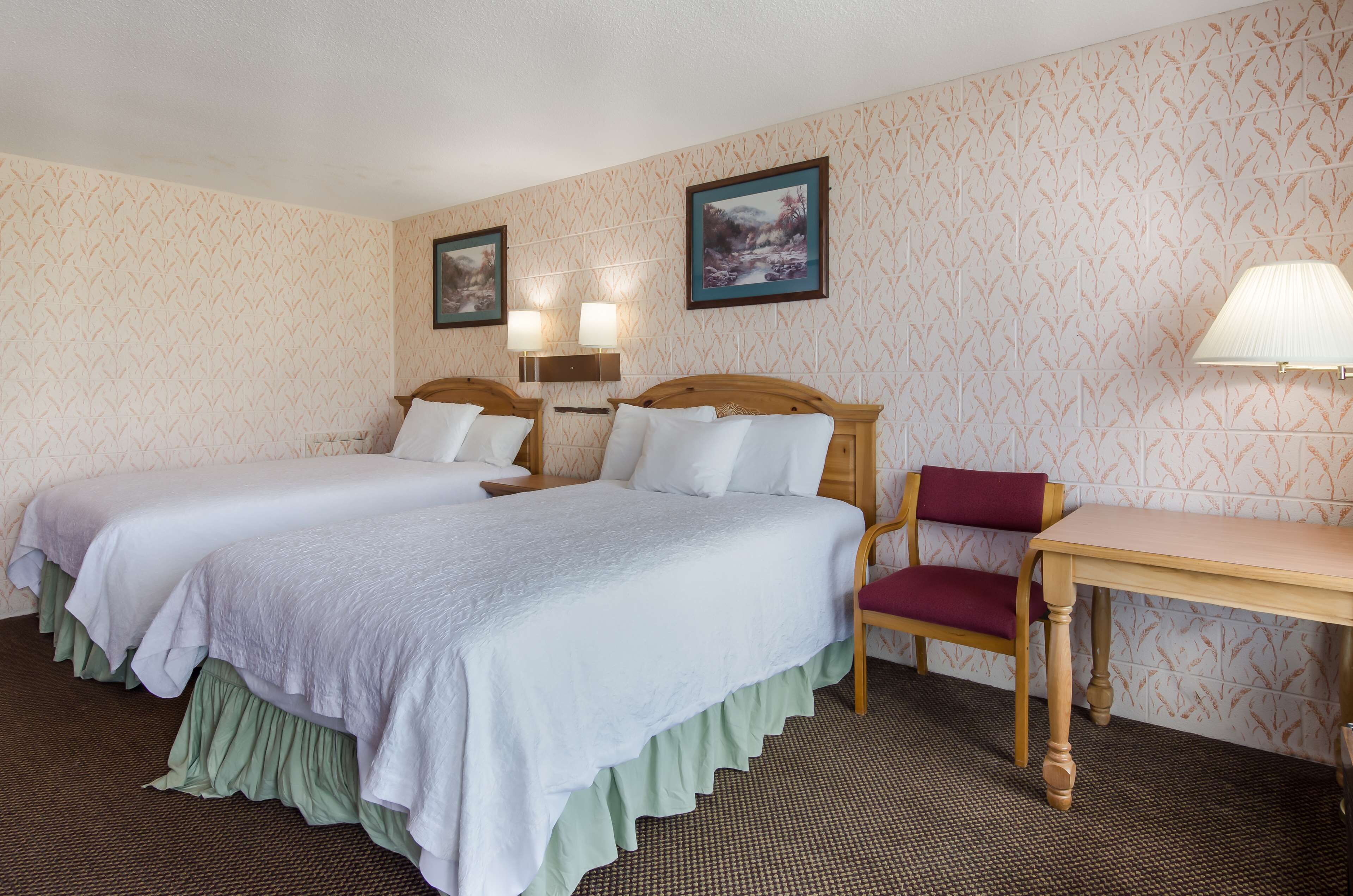 A hotel room with two beds, a wooden table, a chair, and two wall-mounted lamps.