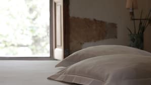 Un lit avec une literie blanche, une fenêtre donnant sur un paysage verdoyant et un mur à la surface texturée.