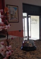 Zona de recepción de un hotel con un cuadro enmarcado de una escena costera, un jarrón de vidrio con flores rosadas y una campana sobre una encimera de mármol.