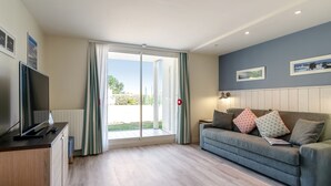A modern living room with a flat-screen TV, a grey sofa with patterned cushions, and a sliding glass door leading to an outdoor area. - Pierre & Vacances Premium Le Domaine de Cramphore (Le Pouliguen)